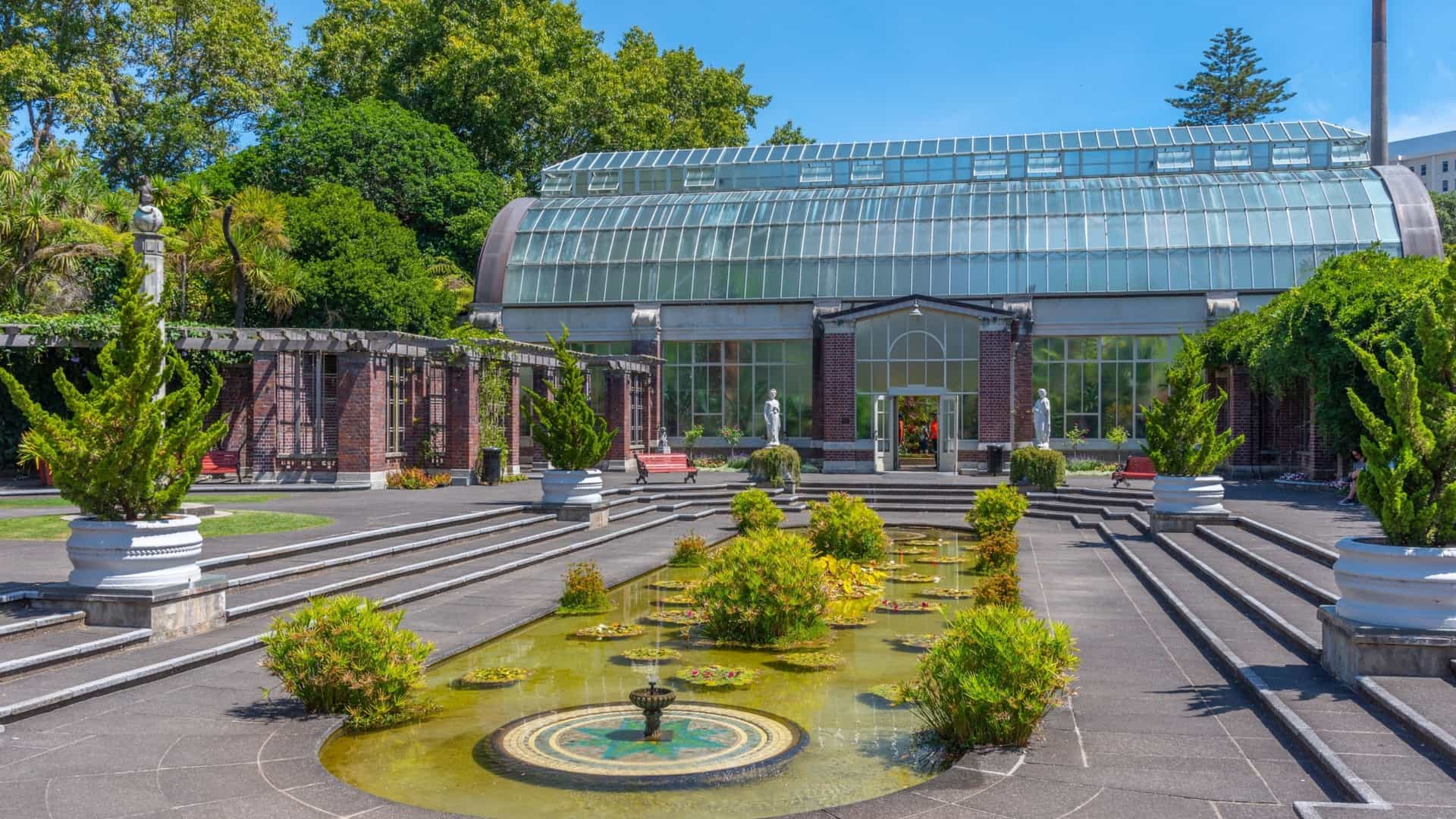 A bright, sunlit view of the Auckland Domain Wintergardens in New Zealand, showing one of the two historic, barrel-vaulted, Victorian-style glasshouses. The heritage glass structure is made of steel and glass, with a curved roof. The foreground features a portion of the ornate, formal courtyard, including a neoclassical marble statue next to a sunken pool with water lilies.
