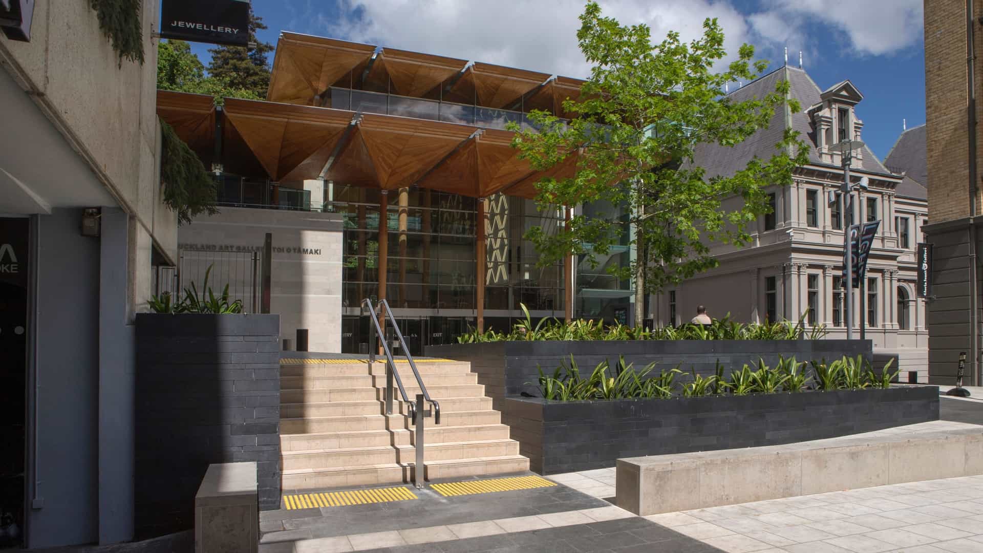 A detail shot of the modern entrance to the Auckland Art Gallery Toi o Tāmaki in New Zealand. The image highlights the distinctive architectural feature of the entrance: a high canopy or roof supported by a series of tall, majestic columns shaped like the branching form of kauri trees. These wooden columns frame the contemporary glass and steel structure of the building.