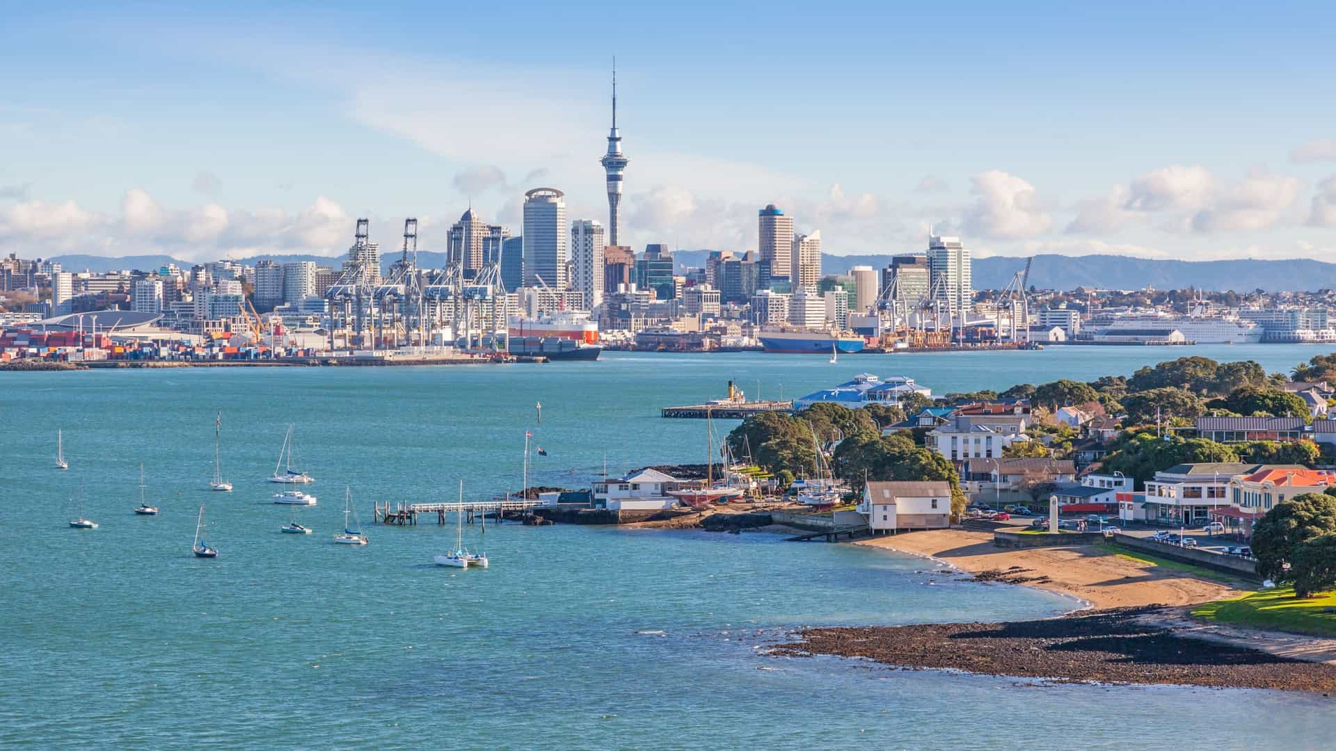 A clear, sunny, panoramic view of the Auckland city skyline and Waitematā Harbour, as seen from the suburb of Devonport in New Zealand. This iconic view is commonly enjoyed after a 12-minute ferry ride to Devonport, Auckland. The foreground features the harbor waters, with a dock extending from the shore, while the background showcases the dense central business district (CBD) skyline, dominated by the distinctive Sky Tower and the Auckland Harbour Bridge spanning the water.