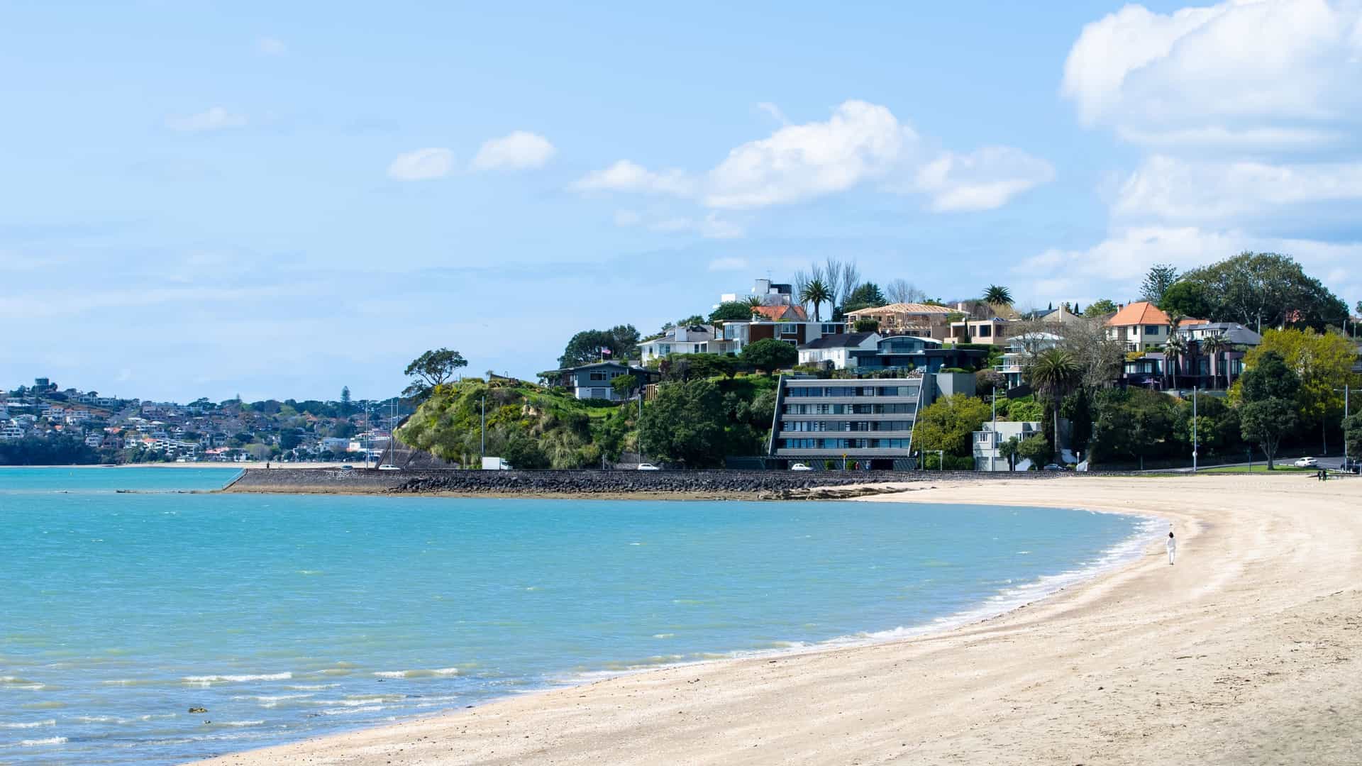 A scenic view of Mission Bay in Auckland, New Zealand, featuring a wide, white-sand beach. In the background, there is a body of water with a small island visible in the distance under a clear sky. The shoreline is lined with a few trees and a path, and there are some people walking along the beach.