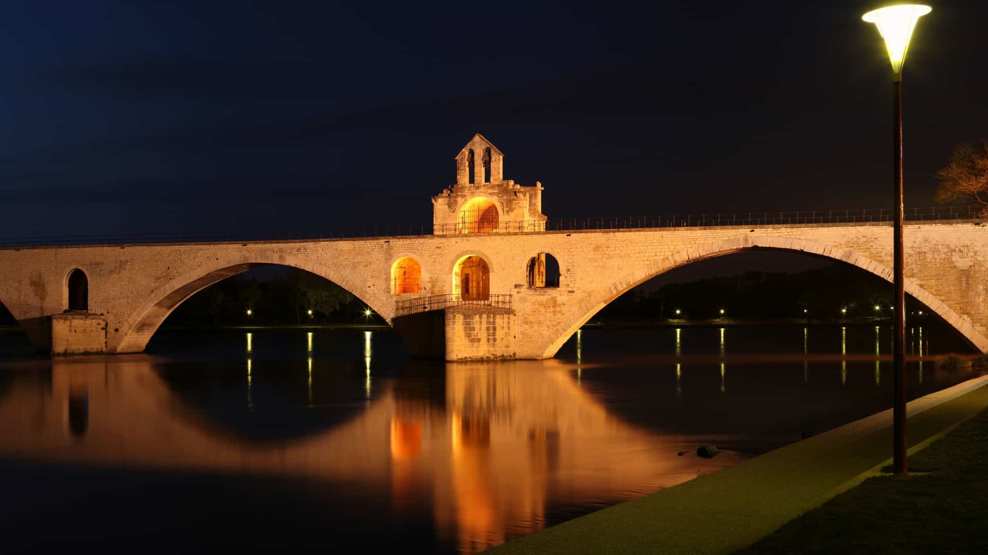 A night view of the illuminated Pont Saint-Bénezet (Pont d'Avignon), a historic medieval bridge over the Rhône River, with the lights of Avignon, France, and the impressive Palace of the Popes visible in the background. The bridge and city lights reflect on the calm surface of the river.