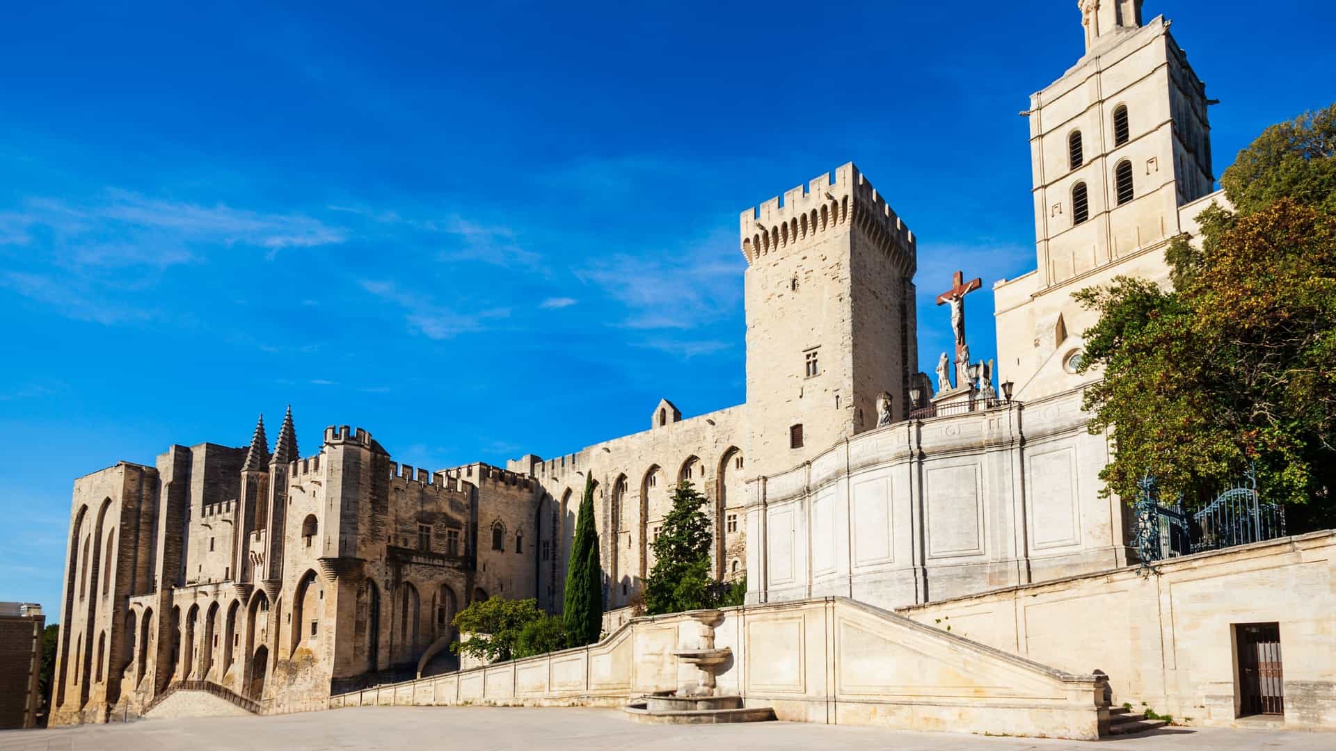 A daytime photo of the Palace of the Popes (Palais des Papes) and the Avignon Cathedral. The massive, fortress-like Gothic palace with its high walls and towers dominates the view, with the smaller cathedral and its golden statue of the Virgin Mary visible next to it. The sky is bright blue.