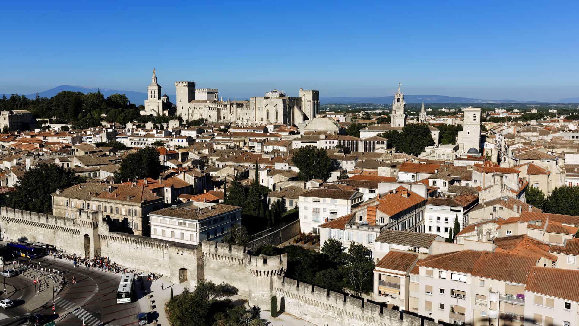 Aerial panoramic view of the historic walled city of Avignon, France, under a clear blue sky. In the center, the dominant landmark is the massive, fortified Palais des Papes (Popes' Palace). The adjacent, slender bell tower of the City Hall is also visible; the square beneath this tower is the Place de l'Horloge, Avignon's central plaza. The foreground features the city's well-preserved medieval ramparts and surrounding urban areas.