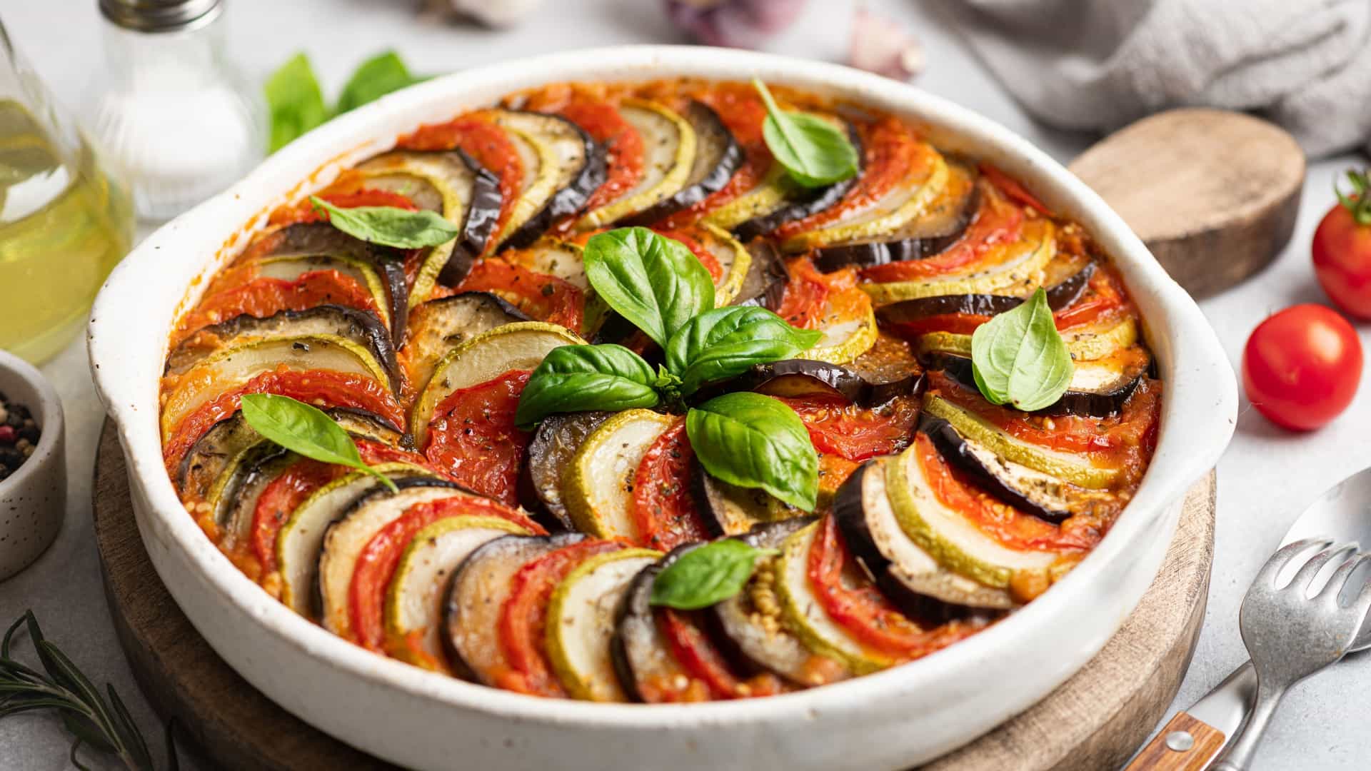 A close-up, high-angle view of ratatouille, a traditional French Provençal vegetable dish. The vegetables—sliced zucchini, yellow squash, tomatoes, and eggplant—are thinly cut and meticulously arranged in an overlapping circular pattern, filling a round white serving dish. The dish is topped with herbs and a drizzle of olive oil, suggesting it has been baked or simmered, with a shallow depth of field that highlights the textures and vibrant colors of the vegetables.
