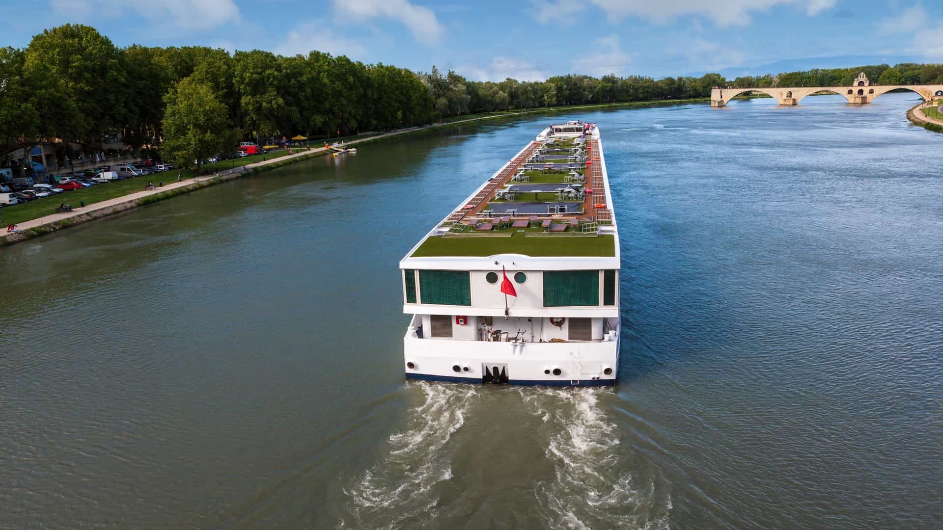 A white, modern river cruise ship, known as a longship, navigates the broad, calm waters of the Rhône River in France. The ship is viewed from the riverbank, with a green shoreline or a distant cityscape visible under a clear sky.