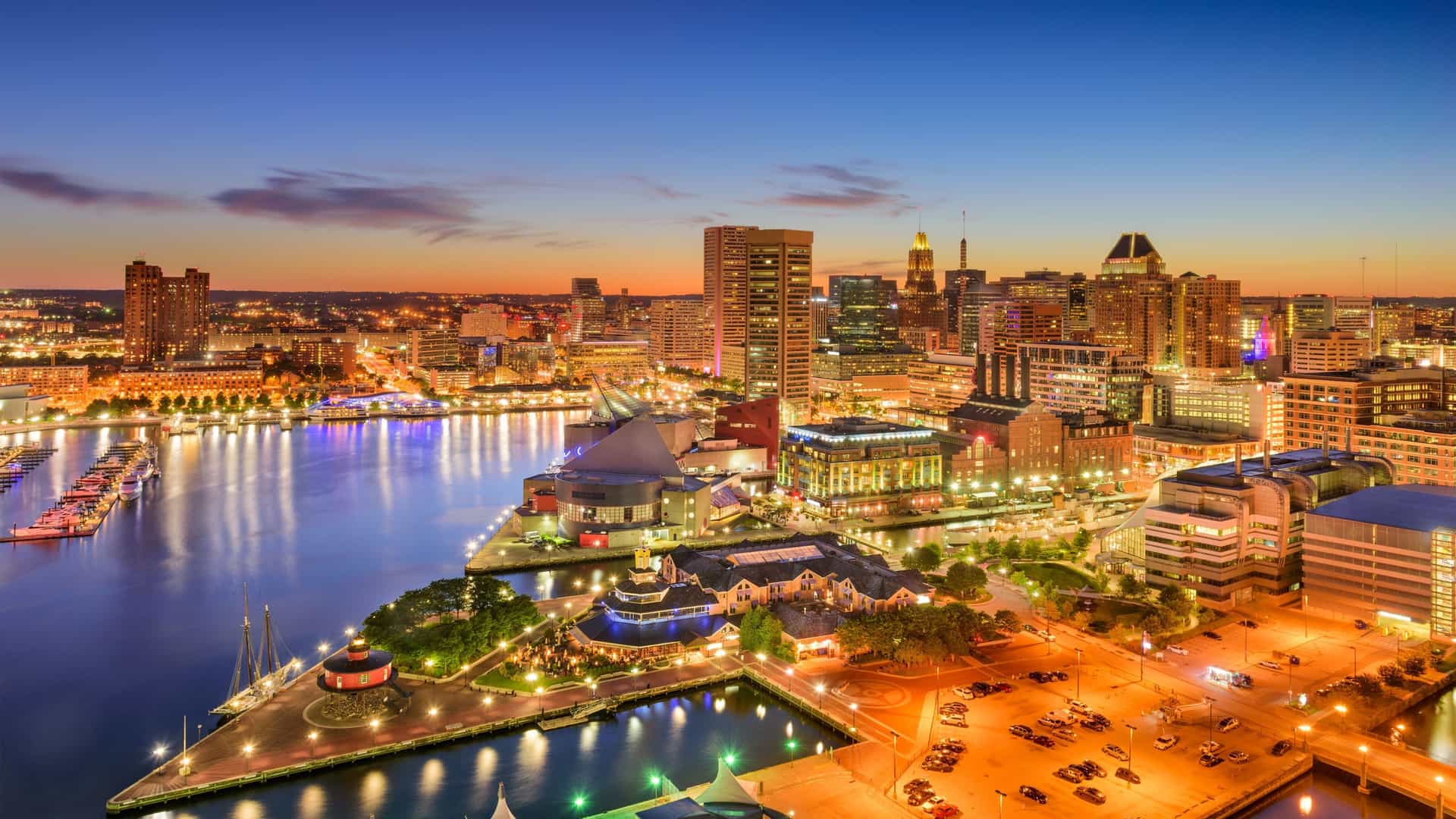 A twilight photo of the Baltimore Inner Harbor with the city's downtown skyline visible in the background. The water reflects the lights of the buildings, and boats are visible docked in the harbor.
