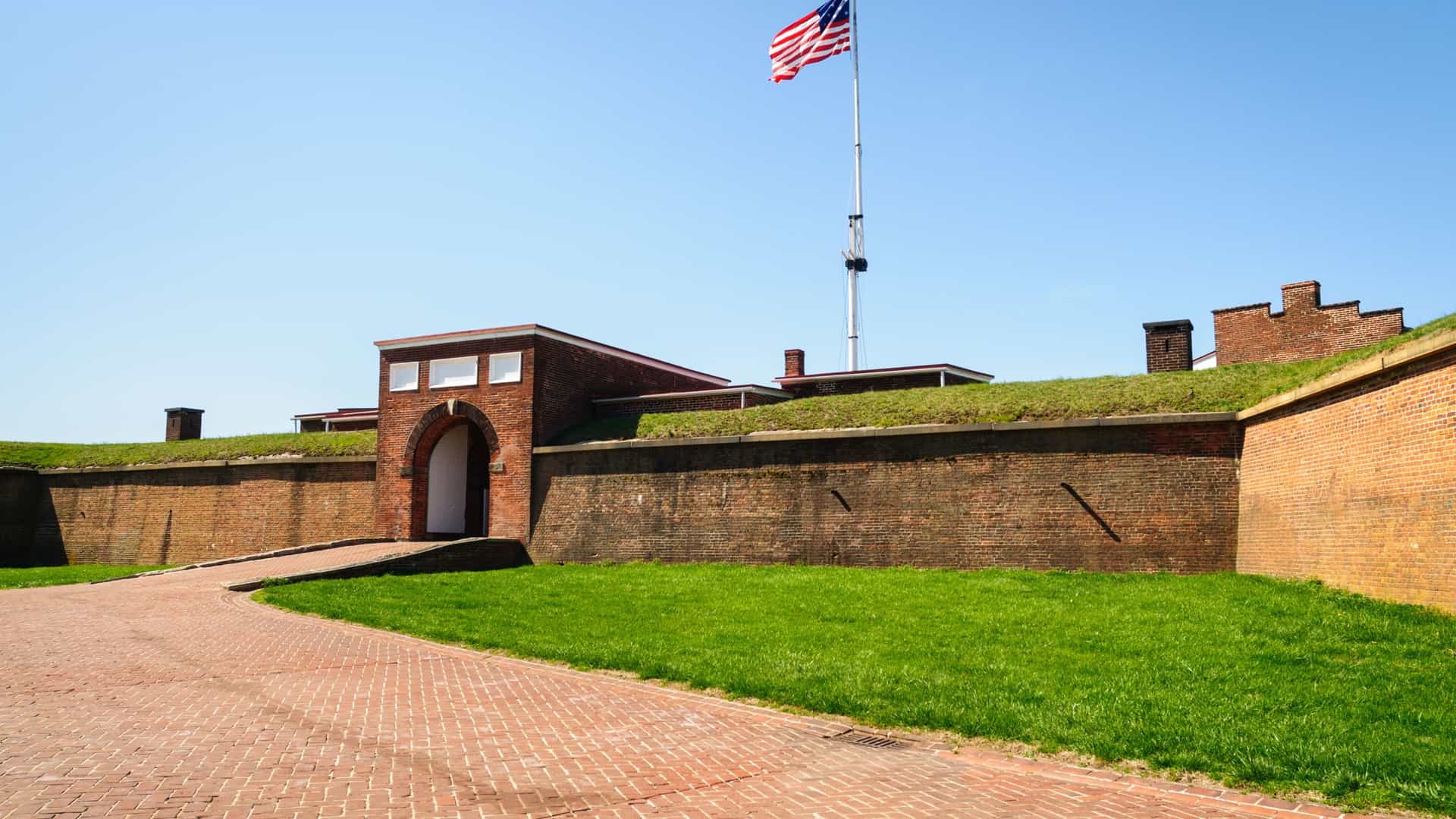 A high-angle view of Fort McHenry, a star-shaped fort on the Baltimore Harbor in Maryland. The fort's red brick and grassy grounds are visible, with a large American flag flying from a flagpole in the center. The surrounding water and a portion of the city's waterfront are in the background.