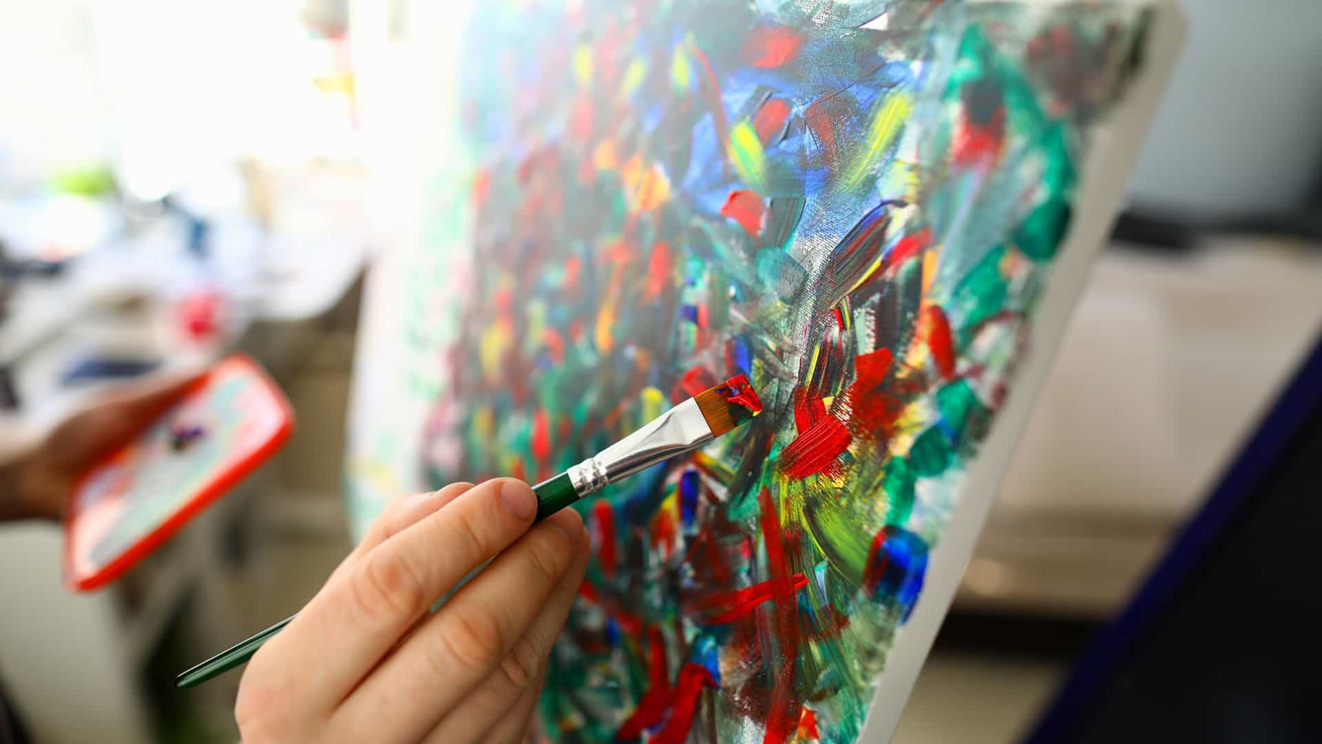 A close-up, selective focus view of an artist's hand using a paintbrush with green handle to apply thick, vibrant red and orange paint to an abstract, colorful canvas on an easel. The painting is a gestural abstract work in progress with visible, thick impasto strokes of red, blue, green, and yellow. A paint palette is visible in the background being held by the artist's other hand.