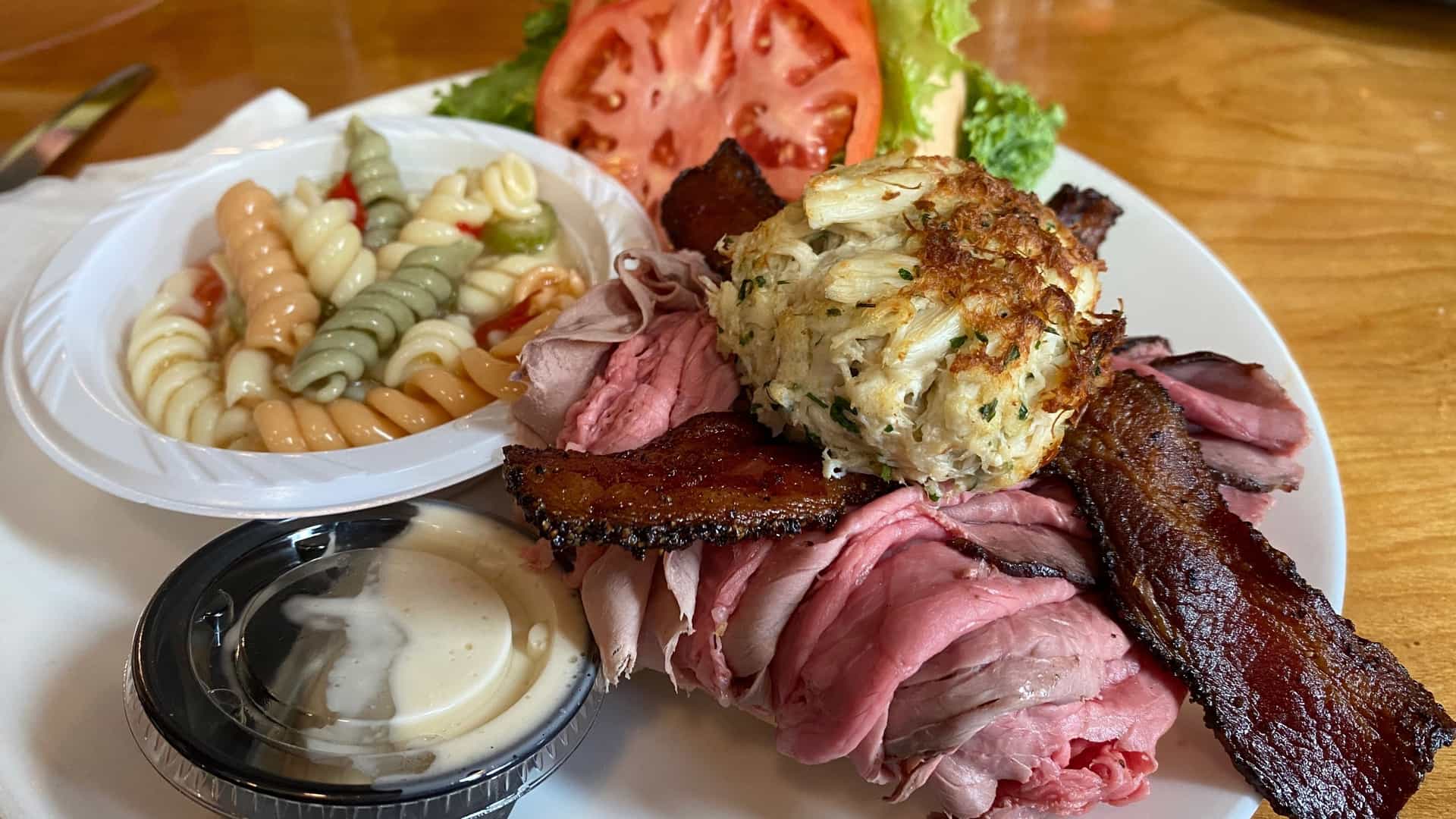 A close-up view of a plate containing two classic Maryland dishes. On the left is a pit beef sandwich, featuring thinly-sliced, dark, grilled roast beef piled high on a roll, likely topped with onions and a white horseradish sauce. On the right is a large, golden-brown, baked or broiled crab cake.
