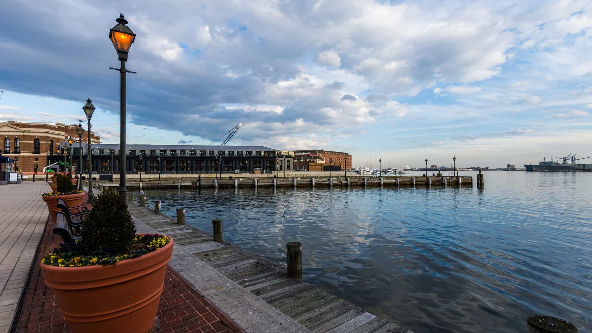 A scenic, long-exposure night shot of the historic Fells Point neighborhood waterfront in Baltimore, Maryland. The view captures the lights of the low-rise 18th and 19th-century brick buildings along the cobblestone (Belgian block) Thames Street, with the glowing reflections of the lights stretching across the dark, still water of the harbor.