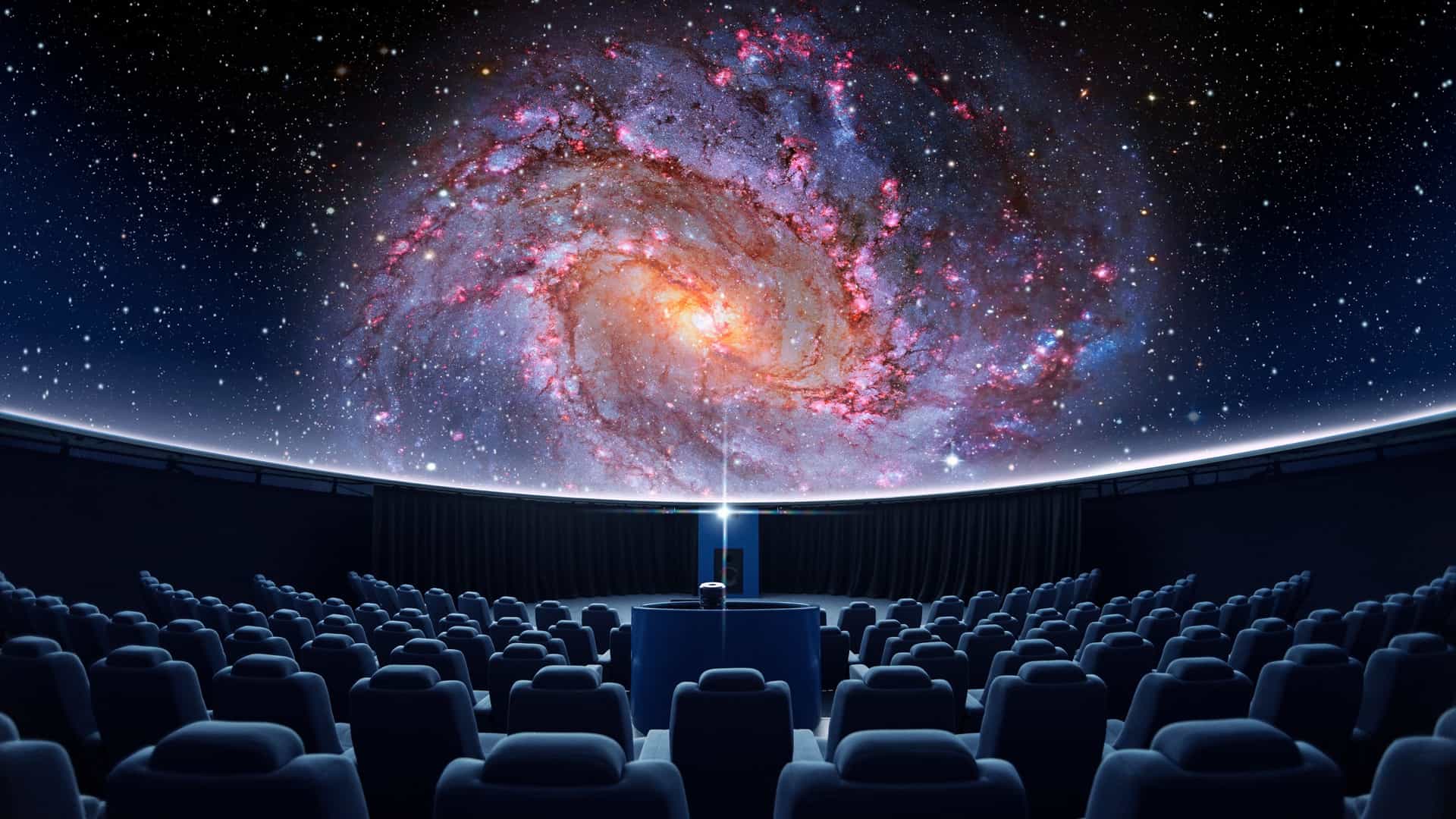 A view inside a planetarium with rows of empty seats in the foreground, facing a massive, curved dome screen that displays a spectacular digital projection of a galaxy, featuring swirling stars and bright cosmic clouds.