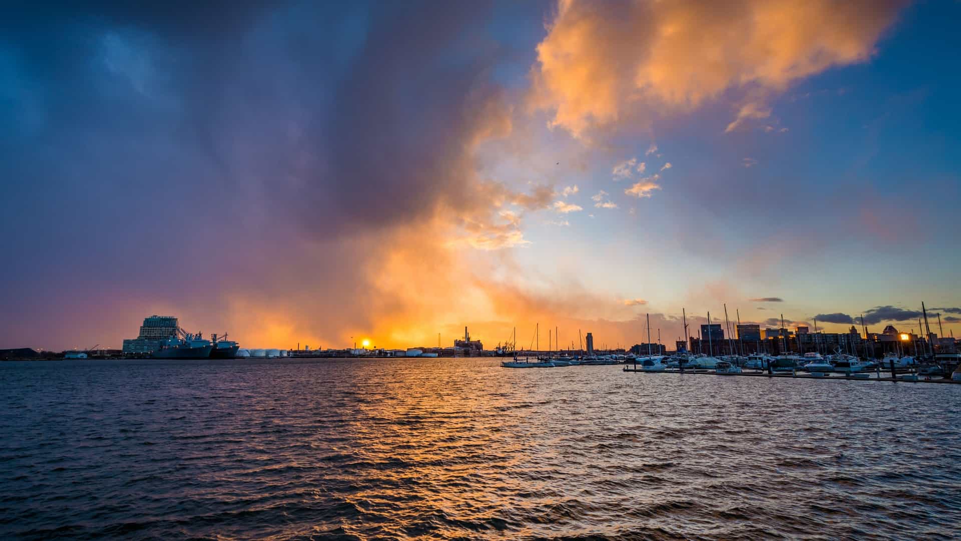 A dramatic sunset over the Patapsco River in Baltimore, Maryland, specifically looking out from the Canton waterfront. The sky is filled with vibrant orange, pink, and yellow hues, with the light reflecting strongly off the water. The silhouette of a distant boat or ship is visible on the water, and the buildings of the Canton or neighboring harbor areas line the shore in the foreground.