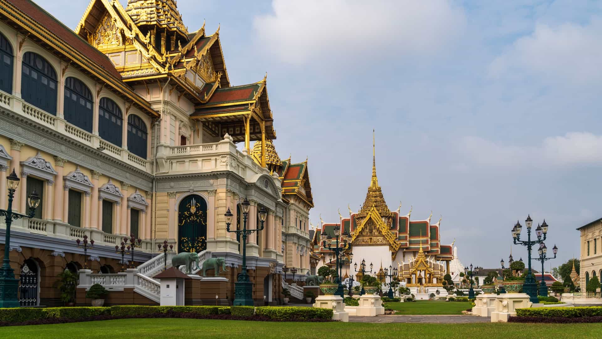 The magnificent, heavily gilded and multicolored architecture of the Temple of the Emerald Buddha (Wat Phra Kaew) complex within the Grand Palace in Bangkok, Thailand, featuring towering chedis, intricate spires, and vibrant mosaics under a blue sky.