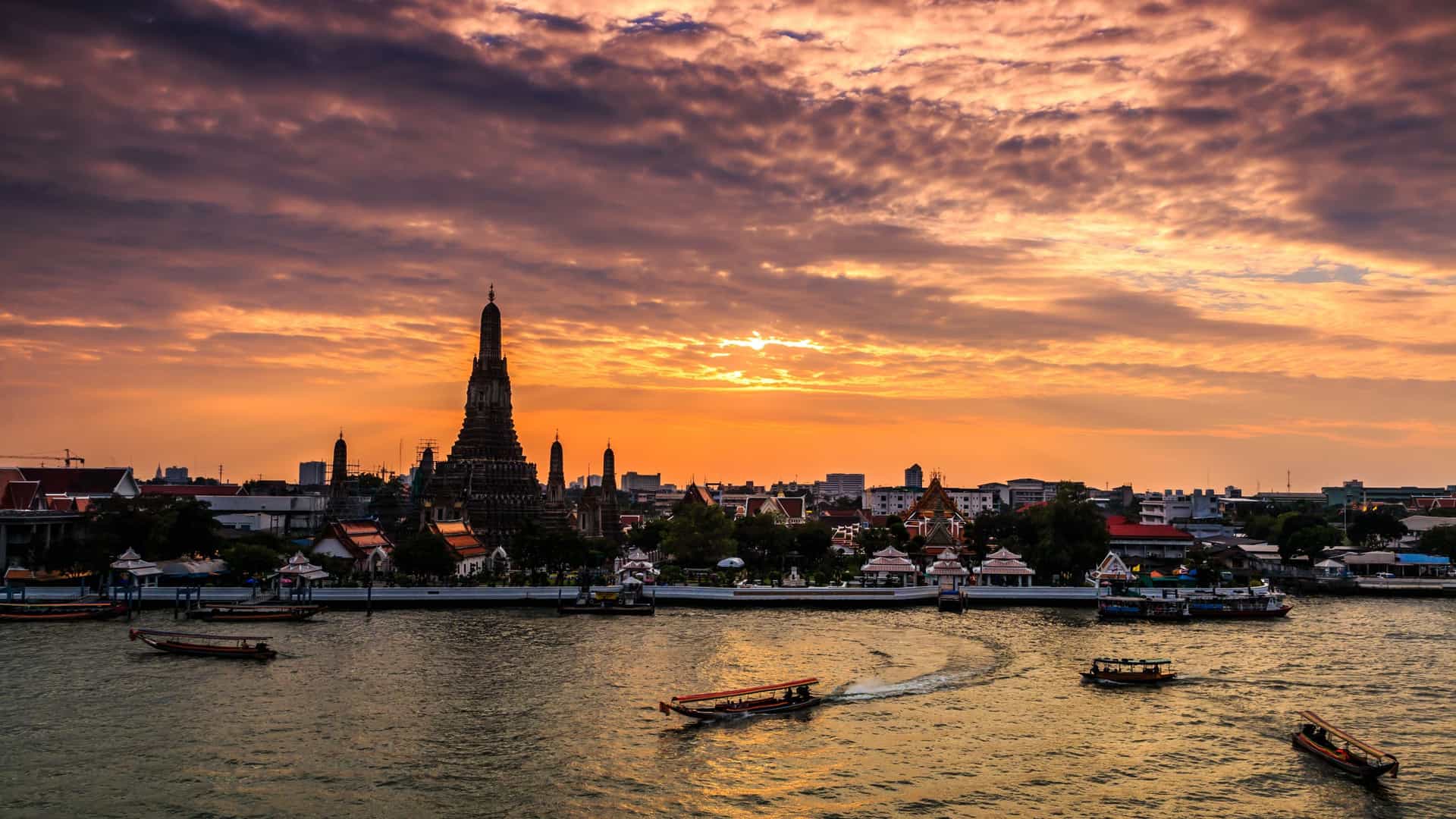 A dramatic sunset over the Chao Phraya River in Bangkok, with the iconic central spire (prang) of Wat Arun (the Temple of Dawn) silhouetted against a brilliant orange and purple sky. Several long-tail boats are visible on the water, suggesting people crossing the river to visit the temple.