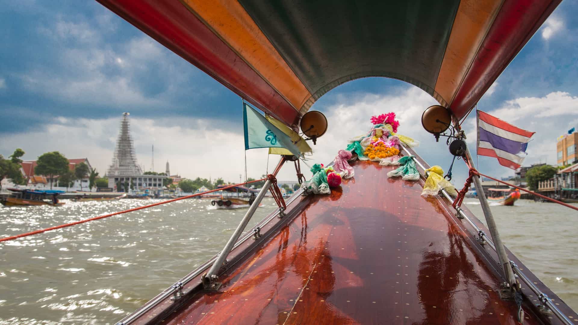 A traditional wooden long-tail boat navigates a narrow, brownish-green canal (khlong) in Bangkok, Thailand. The boat carries several passengers, offering a glimpse into local life along the waterways, which are lined with wooden houses, often built on stilts.