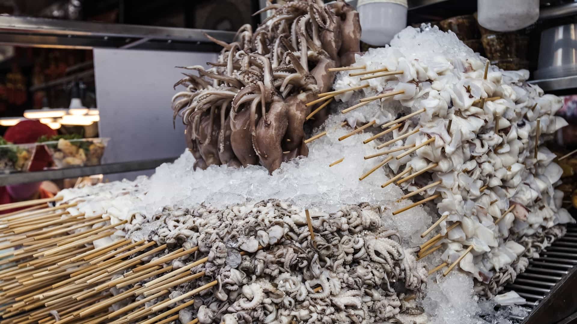 A lively night scene at a seafood barbecue stall in Bangkok's Chinatown (Yaowarat) market. The stall features an abundant display of fresh seafood, including large prawns, crabs, and fish, laid out on ice or skewers, ready to be grilled over hot coals. A vendor is actively cooking or preparing the food, illuminated by the bright market lights.