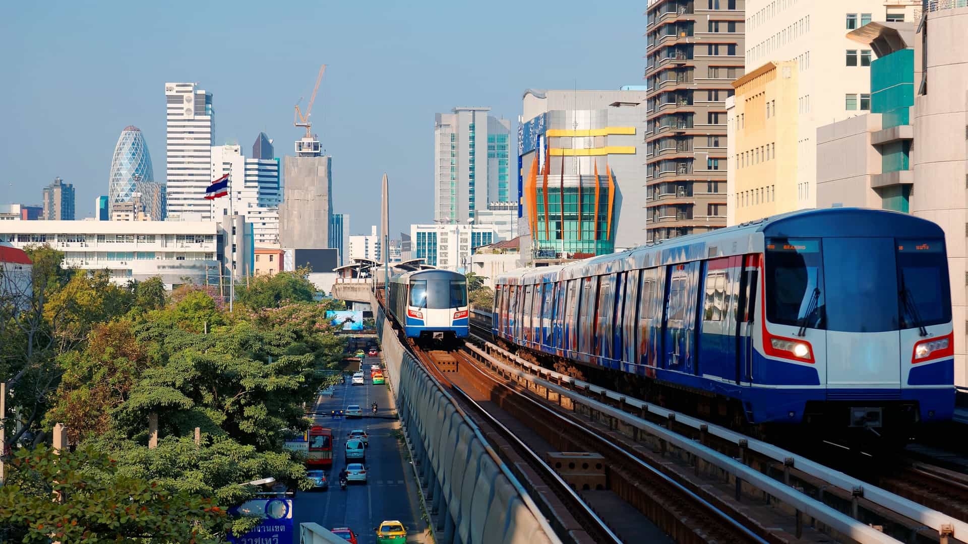 A high-angle daytime cityscape of Downtown Bangkok, Thailand, showcasing a dense array of modern high-rise buildings and skyscrapers, representing the city's fast development. In the foreground, the elevated tracks of the BTS Skytrain run through the urban core, with a train visible, and the Victory Monument prominent in the background.