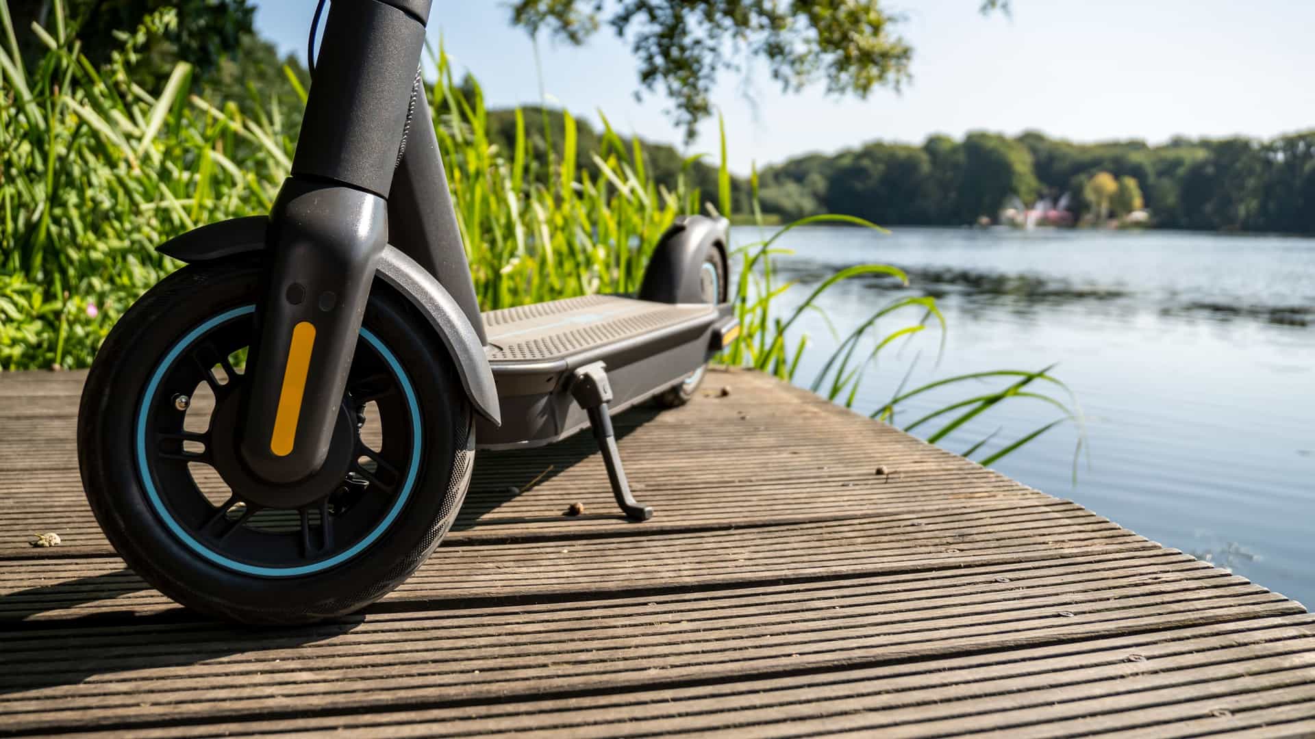 A modern electric kick scooter (e-scooter) is parked on a wooden boardwalk, suggesting a ride along a Malecon. In the background, a calm lake is visible, bordered by green grass and topped by a clear blue sky on a bright, sunny day.