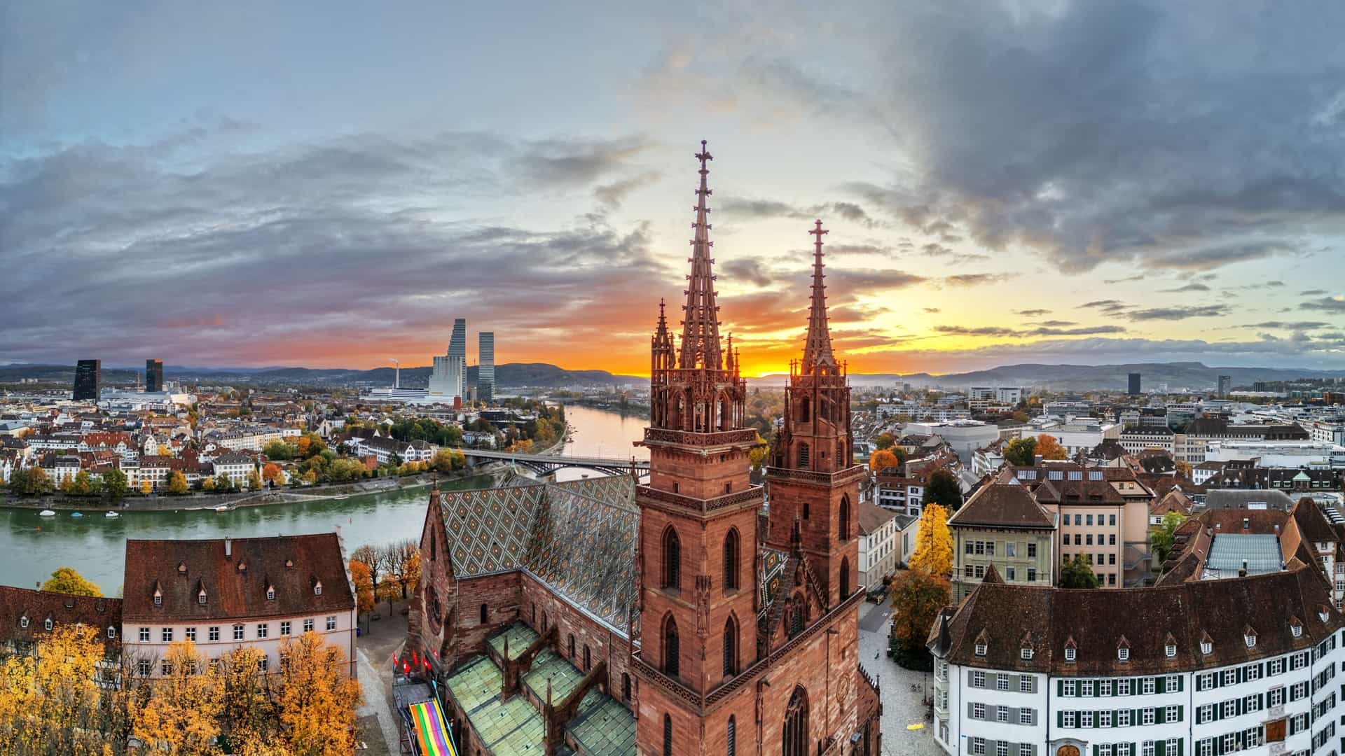 A scenic view of Basel, Switzerland at sunrise, with the Rhine River in the foreground reflecting the early morning light. The historic Old Town, including the Basel Cathedral (Münster) and the Mittlere Brücke (Middle Bridge), is visible on one side of the river, while modern high-rise buildings, such as the Roche Tower, line the opposite bank.