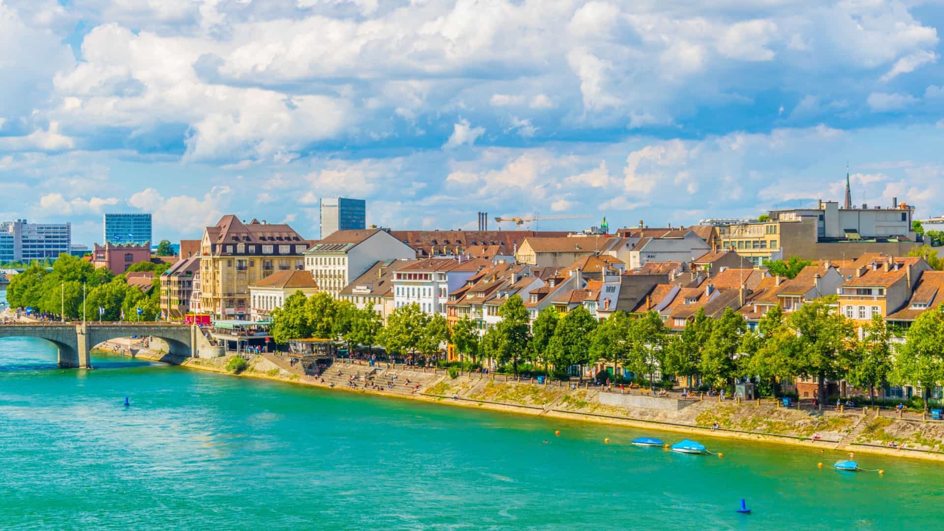 A bright panoramic view of the Rhine River flowing through Basel, Switzerland. On the far bank, the historic city skyline of Grossbasel is dominated by the red sandstone Basler Münster (Cathedral) with its distinctive twin towers, rising above the densely packed, centuries-old buildings lining the riverfront.