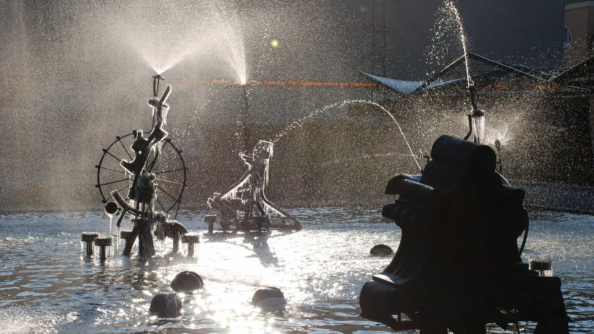 A close-up view of the Tinguely Fountain in Basel, a kinetic art sculpture by Jean Tinguely. The fountain features several large, whimsical black metal machines, constructed from scrap mechanical parts, moving and spraying jets of water into a shallow pool. The historic stone walls of the surrounding buildings and the square are visible in the background.