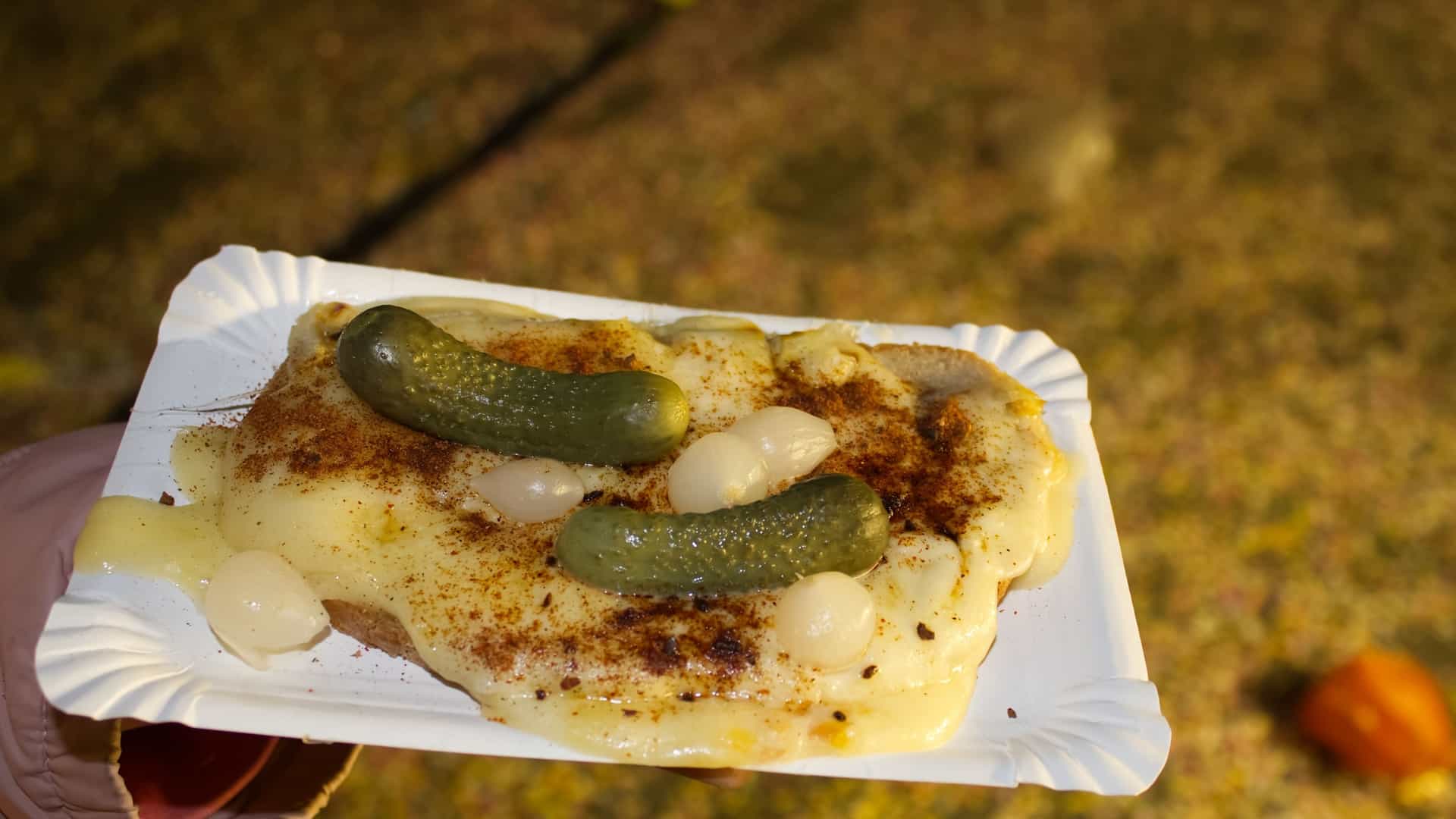 A close-up of a slice of bread generously topped with a layer of melted, gooey Raclette cheese. The cheese is garnished with slices of gherkin pickles and chopped onion. The food is presented on a white paper plate, suggesting it is a street food item from a carnival or market.