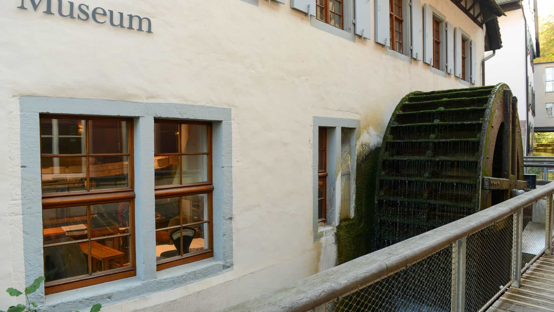 The Paper Mill Museum Basel on Switzerland. A picturesque view of the museum housed in an historic, half-timbered building situated next to a canal with a functioning waterwheel. The building is surrounded by greenery under a blue sky.