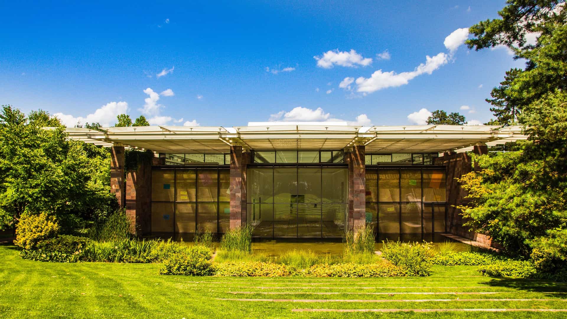 The modern Fondation Beyeler art museum in Riehen, Switzerland, a building designed by architect Renzo Piano. The structure features a long facade of red porphyry stone walls and a distinctive lightweight glass roof, set in a tranquil, manicured park environment.