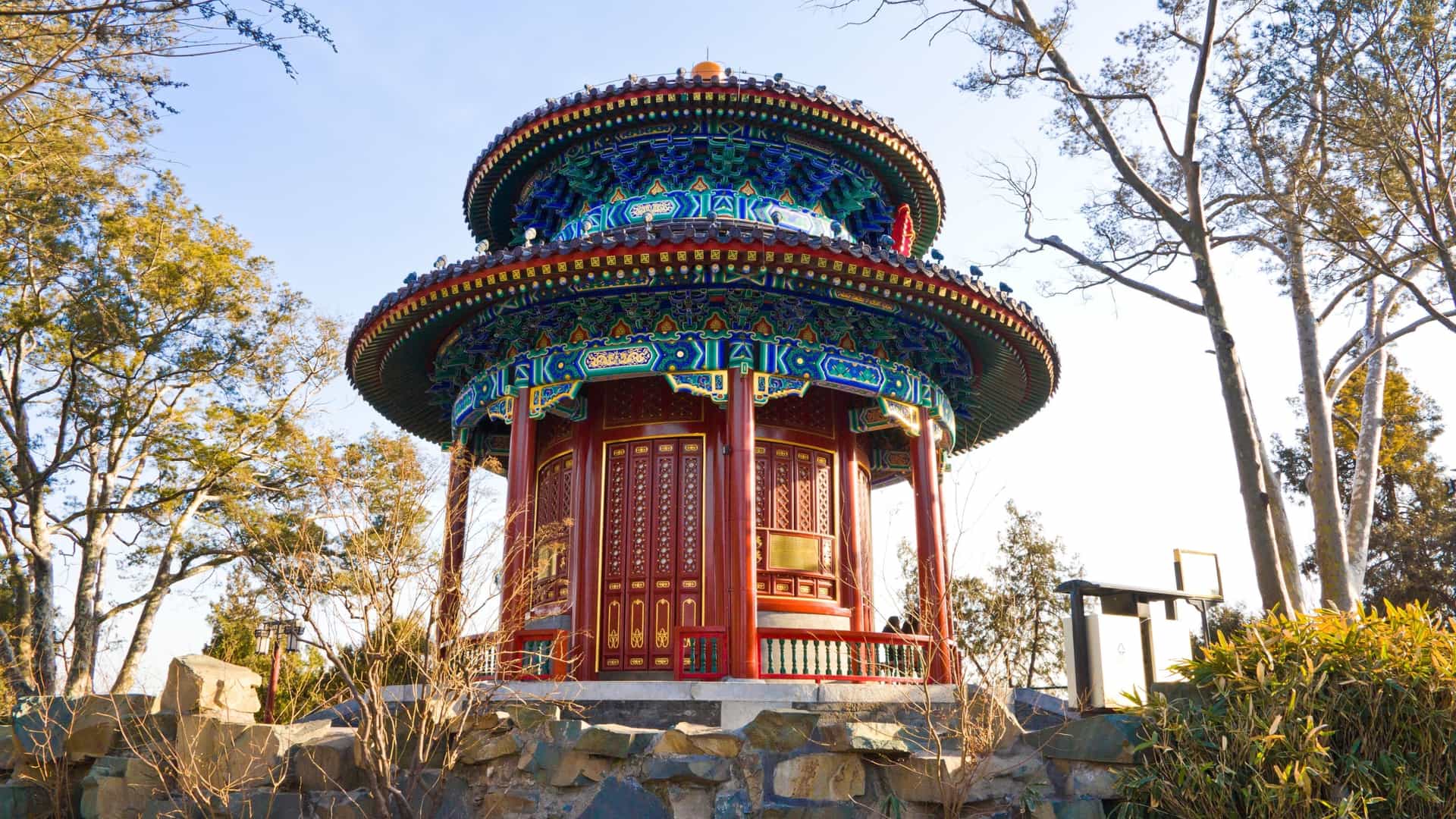 A close up view of the Wanchun Pavilion in Jingshan Park, Beijing, featuring a magnificent, multi tiered traditional Chinese roof with imperial yellow glazed tiles, intricate eaves, and decorative roof ridges, set against a backdrop of trees under a clear sky.