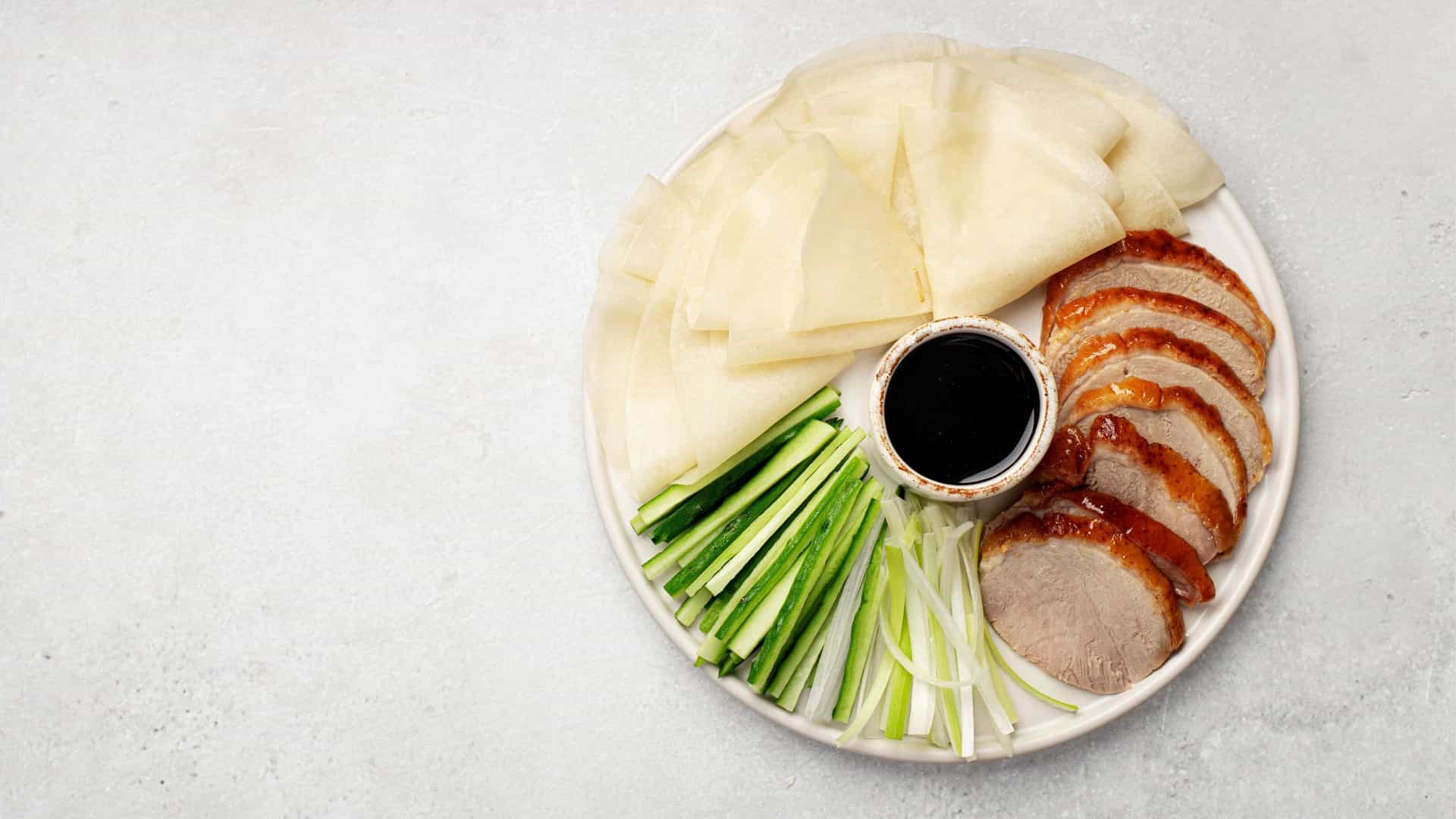 A close up, top down view of a traditional Peking Duck dish. The roasted duck, with its signature crispy, golden brown skin, is artfully sliced and arranged on a white plate, accompanied by a small bowl of hoisin sauce and a side of fresh cucumber and green onion strips.
