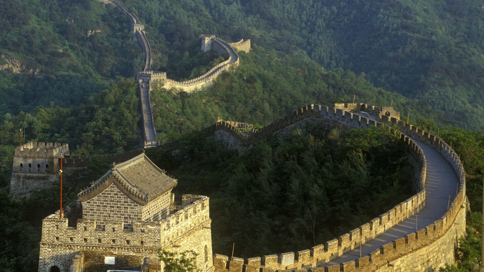 A clear, sunny day view of the Mutianyu section of the Great Wall of China. The wall, with its numerous watchtowers, snakes its way across the verdant, forested mountain ridges, with a small cluster of buildings visible in the valley below.