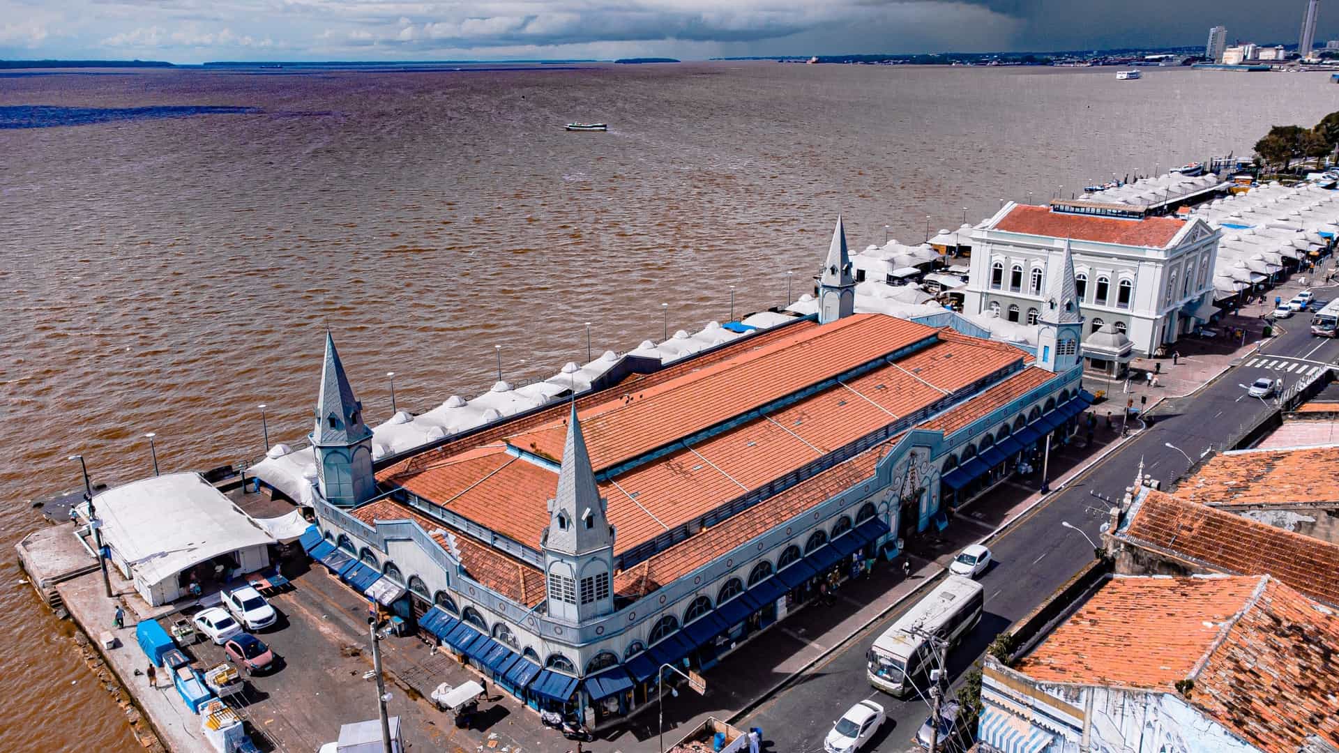 A vibrant, wide angle landscape view of the historic Ver o Peso Market complex in Belém, Pará, situated on the Guajará Bay near the mouth of the Amazon River. The scene features the market's main colonial era buildings, including the distinctive green Iron Market (Mercado de Ferro) structure, alongside a busy waterfront where numerous colorful wooden fishing boats are docked, reflecting the Amazonian commerce.