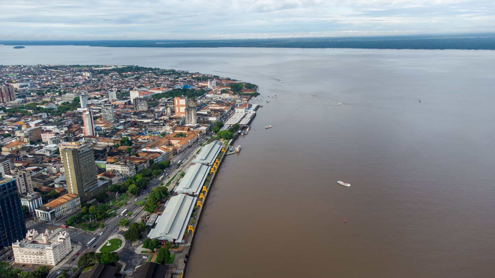 An aerial view of the Estação das Docas, a renovated port area in Belém, Brazil. The complex features three large warehouses with red roofs and a riverside promenade lined with restaurants, shops, and yellow industrial cranes. The area is situated along the banks of the Guajará Bay, with the cityscape of Belém visible in the background.
