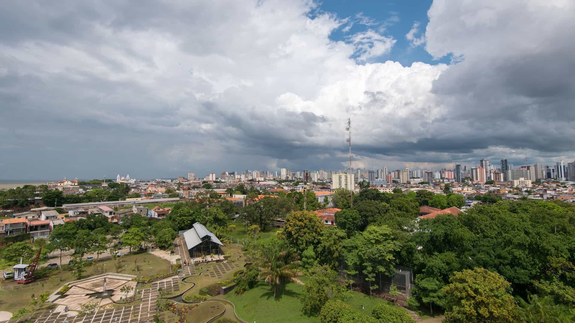 An aerial view of Mangal das Garças Naturalistic Park in Belém, Brazil. The image shows a lush green park with a central tower and landscaped gardens next to the Guamá River. The cityscape of Belém is visible in the distance, with the park acting as an urban oasis.