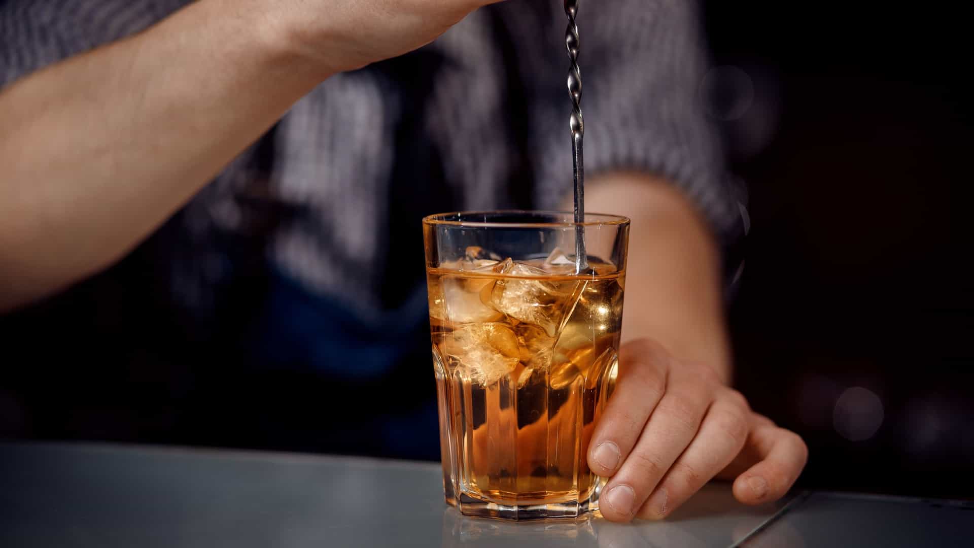 A barman shakes a cocktail in a metal shaker with dramatic energy behind a long, dark wooden bar counter. A clear glass with ice awaits the drink, suggesting a busy or atmospheric setting. The scene evokes the classic Victorian pub experience of the iconic Crown Liquor Saloon in Belfast.