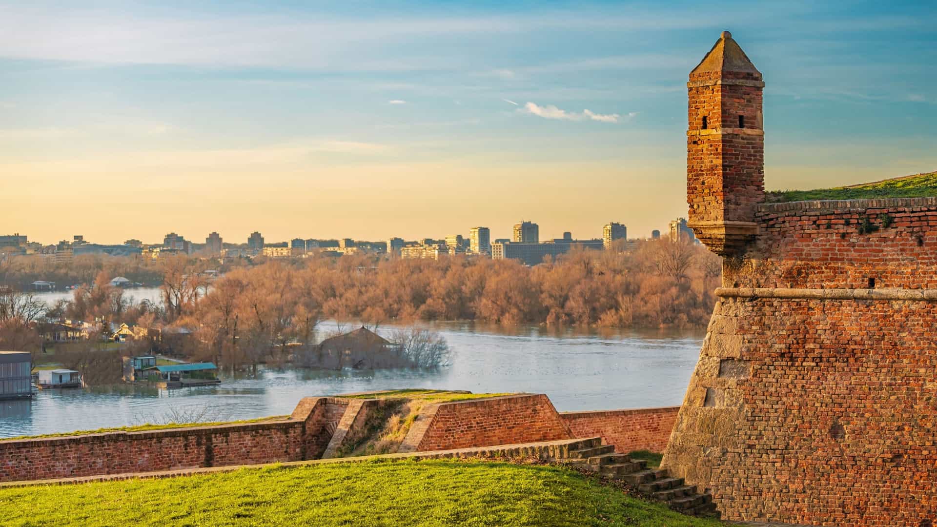 A breathtaking sunset view from Belgrade's Kalemegdan Fortress, with the sky painted in intense golden hues of orange and yellow. In the foreground, the silhouette of the Pobednik (Victor) monument stands tall, overlooking the confluence of the Sava and Danube rivers. The fortified walls of the ancient fortress are visible in the warm, fading light.