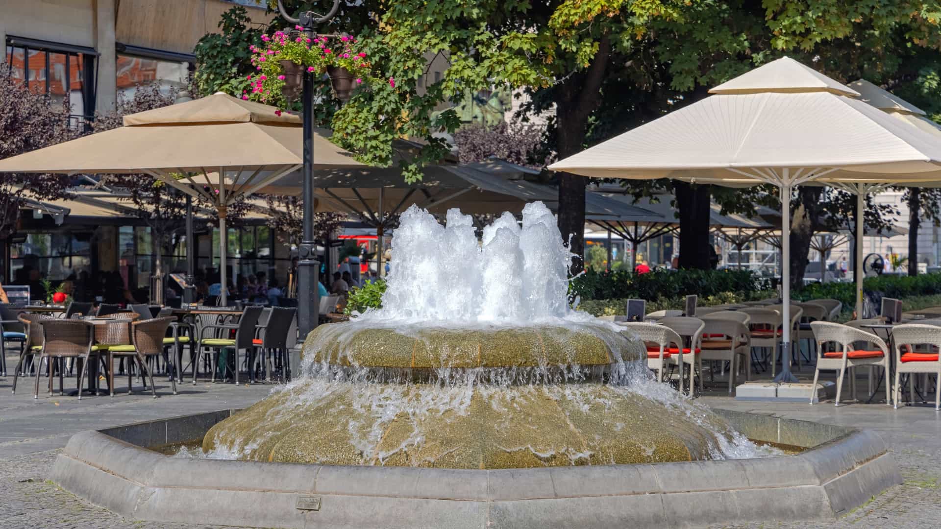 A detail shot of a decorative water fountain on Knez Mihailova Street in Belgrade, Serbia. The fountain features an ornate, classical design, with water gently spilling from a spout into a basin, surrounded by the pedestrian area of the city's main street. The polished stone and bronze elements of the fountain are prominent in the foreground.