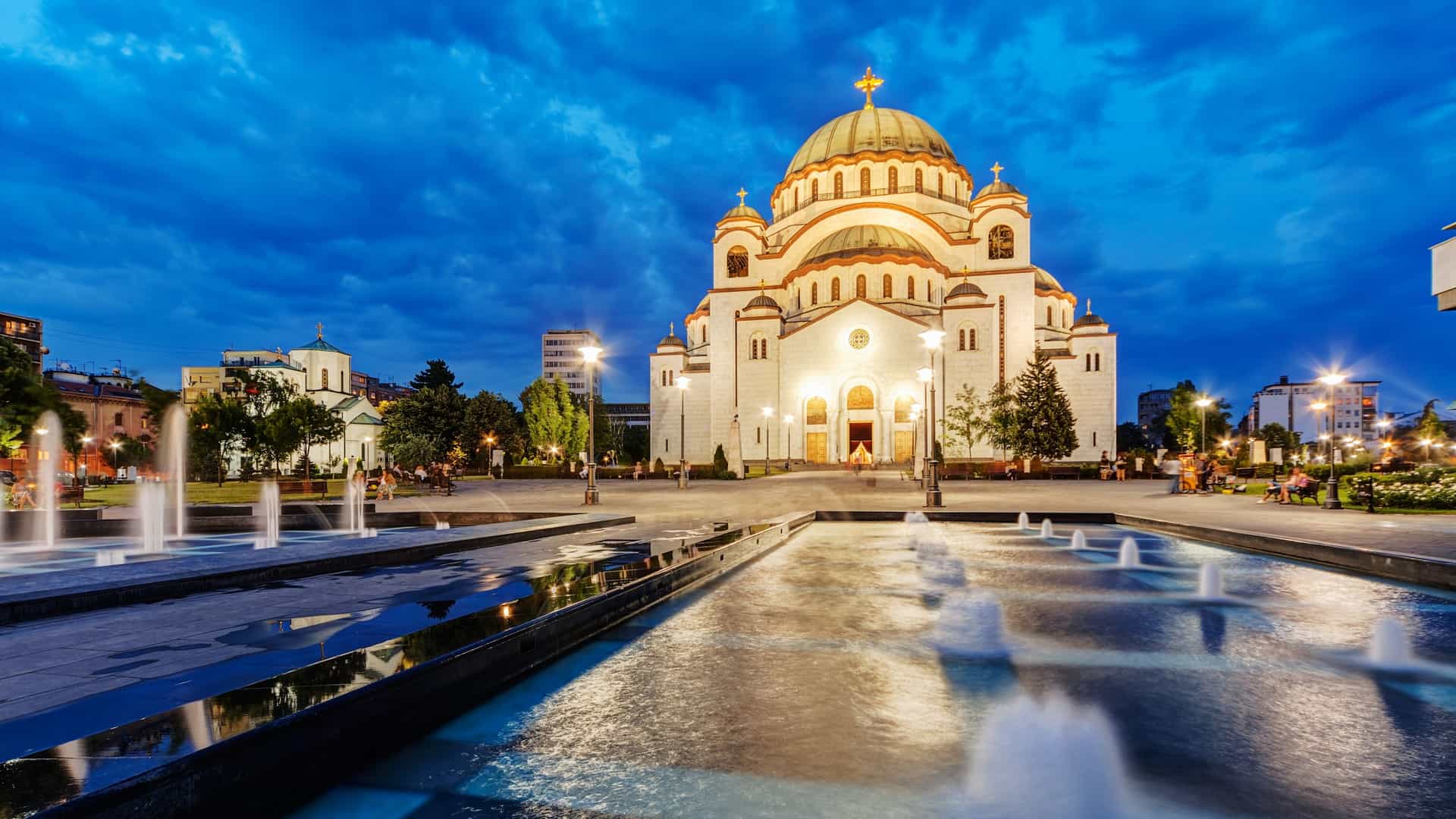 A photo of the Church of Saint Sava in Belgrade, Serbia. The large, white Orthodox church, built in the Byzantine Revival style, is visible with its prominent central dome and multiple smaller domes. The building's exterior is made of white marble and granite, and it is surrounded by a park like setting.