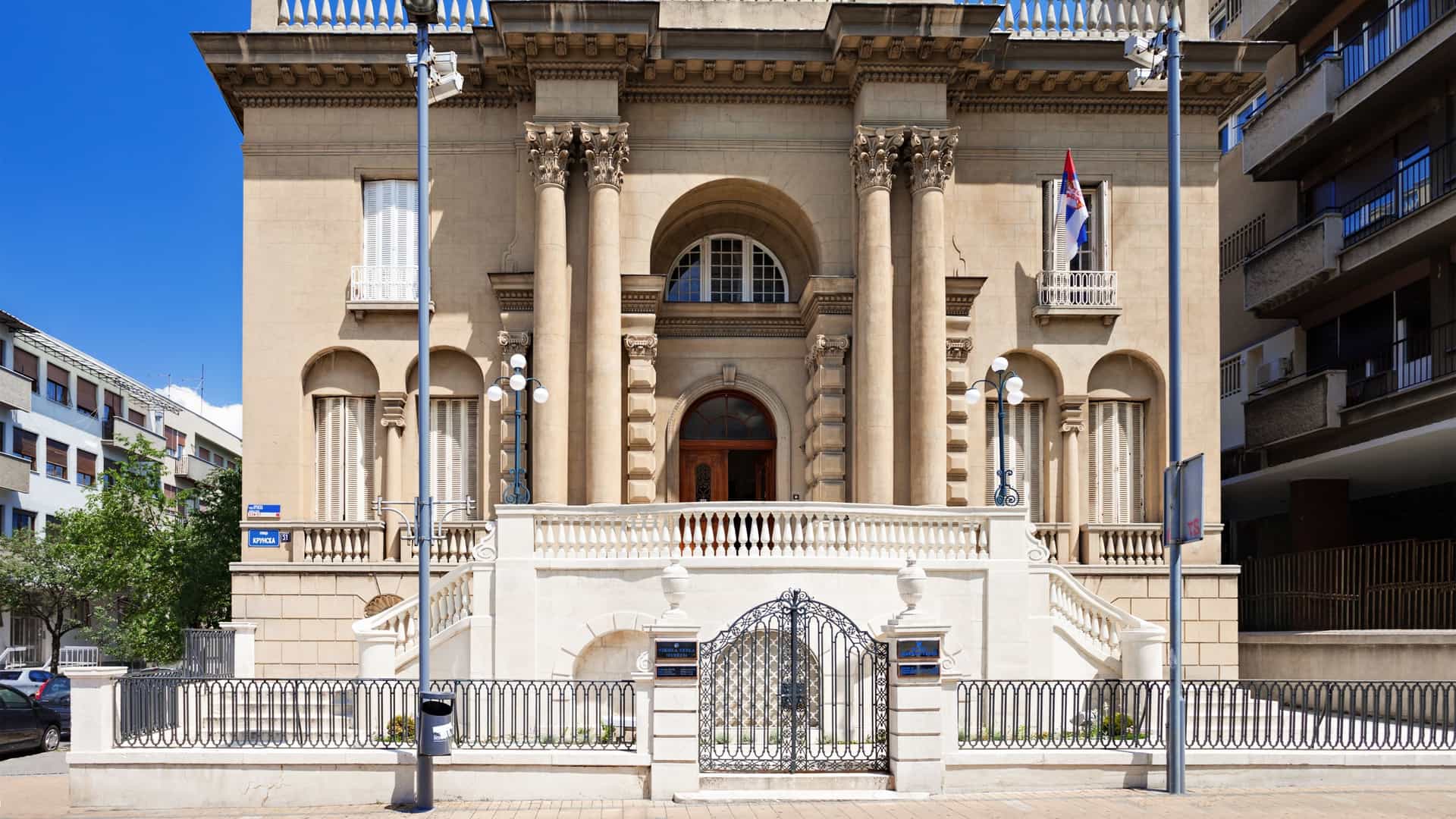 A photo of the Nikola Tesla Museum in Belgrade, Serbia. The museum is housed in a historic villa on Krunska Street, with a classic, stately facade and a small garden area in the front.