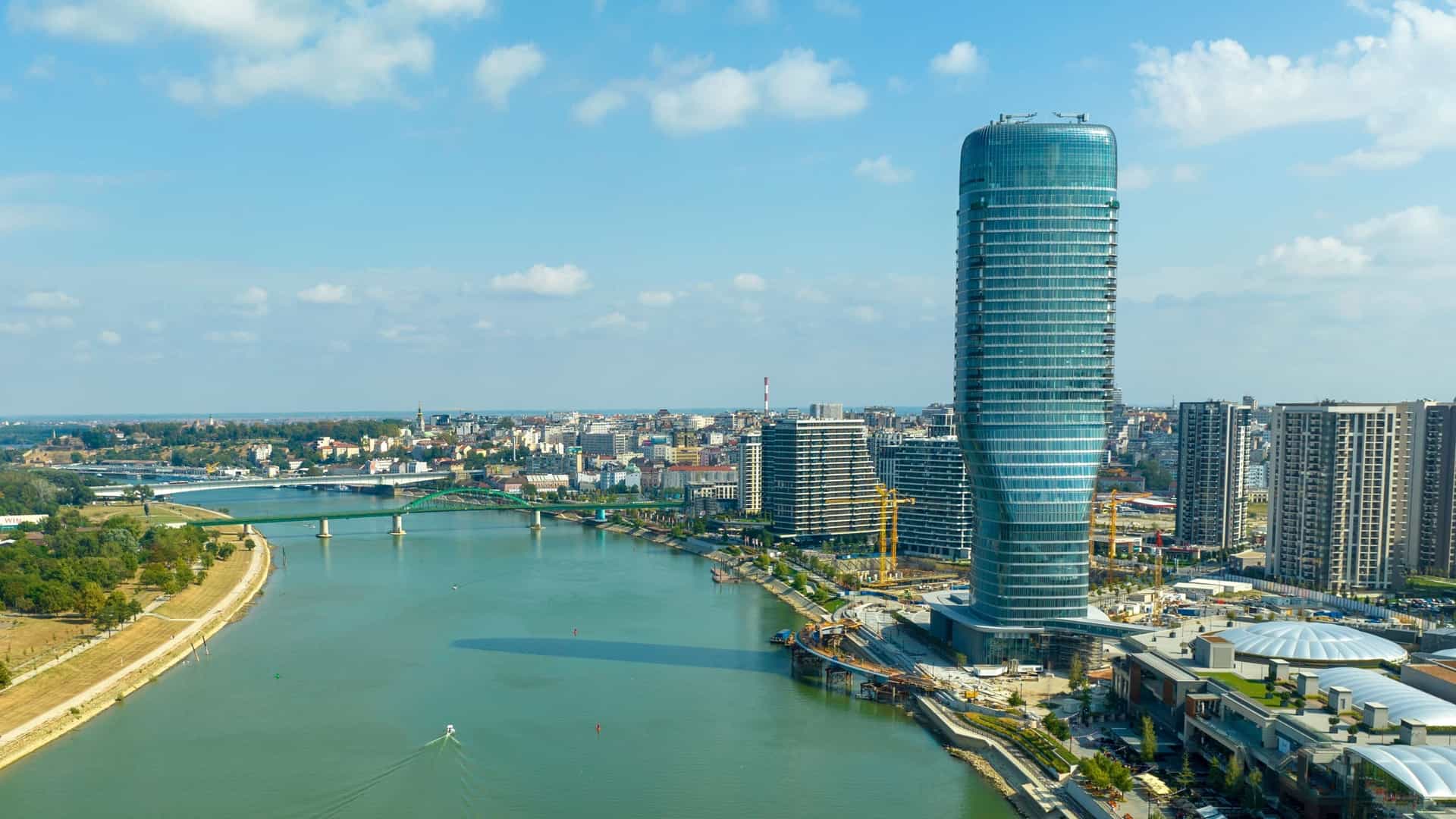 A panoramic, high angle view of the Belgrade Waterfront, a new residential and commercial development, on the banks of the Sava River in Belgrade, Serbia. The river flows through the scene, with modern buildings and bridges visible along its banks.