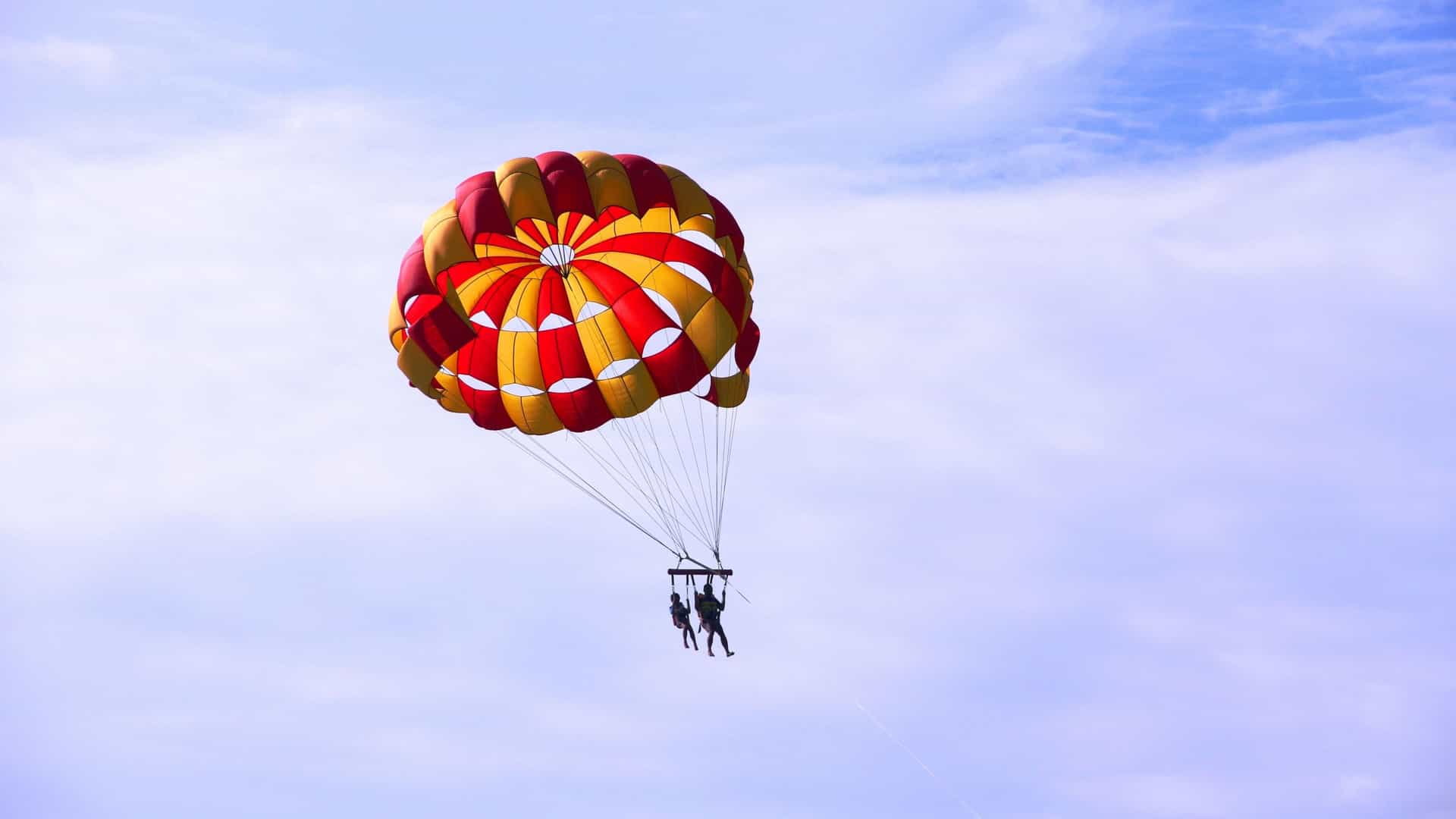 Parasailing over the ocean in a coastal setting, likely Tanjung Benoa, Bali, Indonesia. A person is suspended high in the air by a colorful, parachute like canopy, towed by a speedboat over the turquoise water. The location is famous as Bali's Marine Sports Centre.