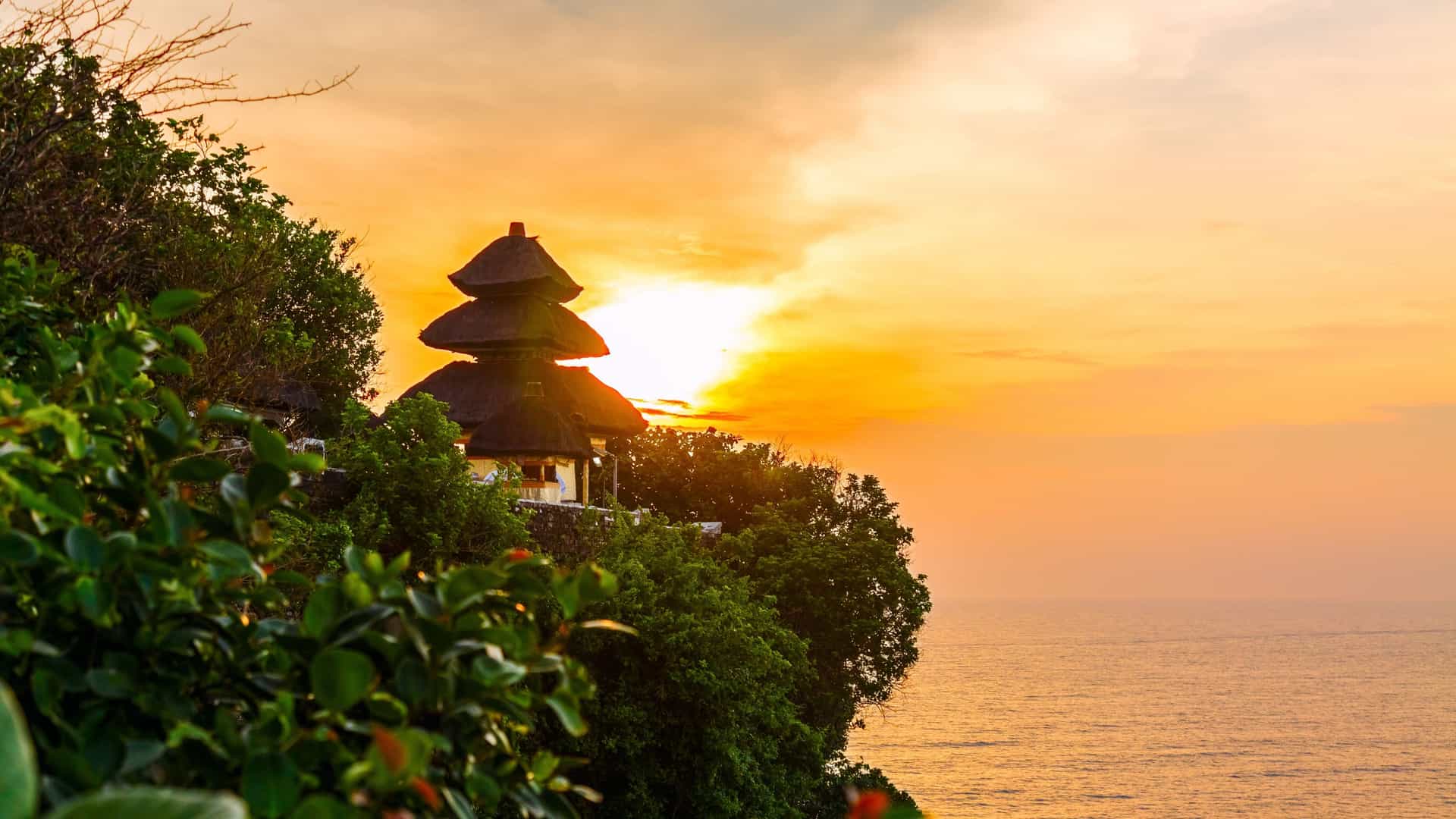 Pura Luhur Uluwatu, a sacred Balinese Hindu sea temple, is silhouetted against a brilliant sunset. The temple is perched dramatically on a steep, rocky cliff overlooking the Indian Ocean, with the sky above a vivid gradient of orange, yellow, and red.