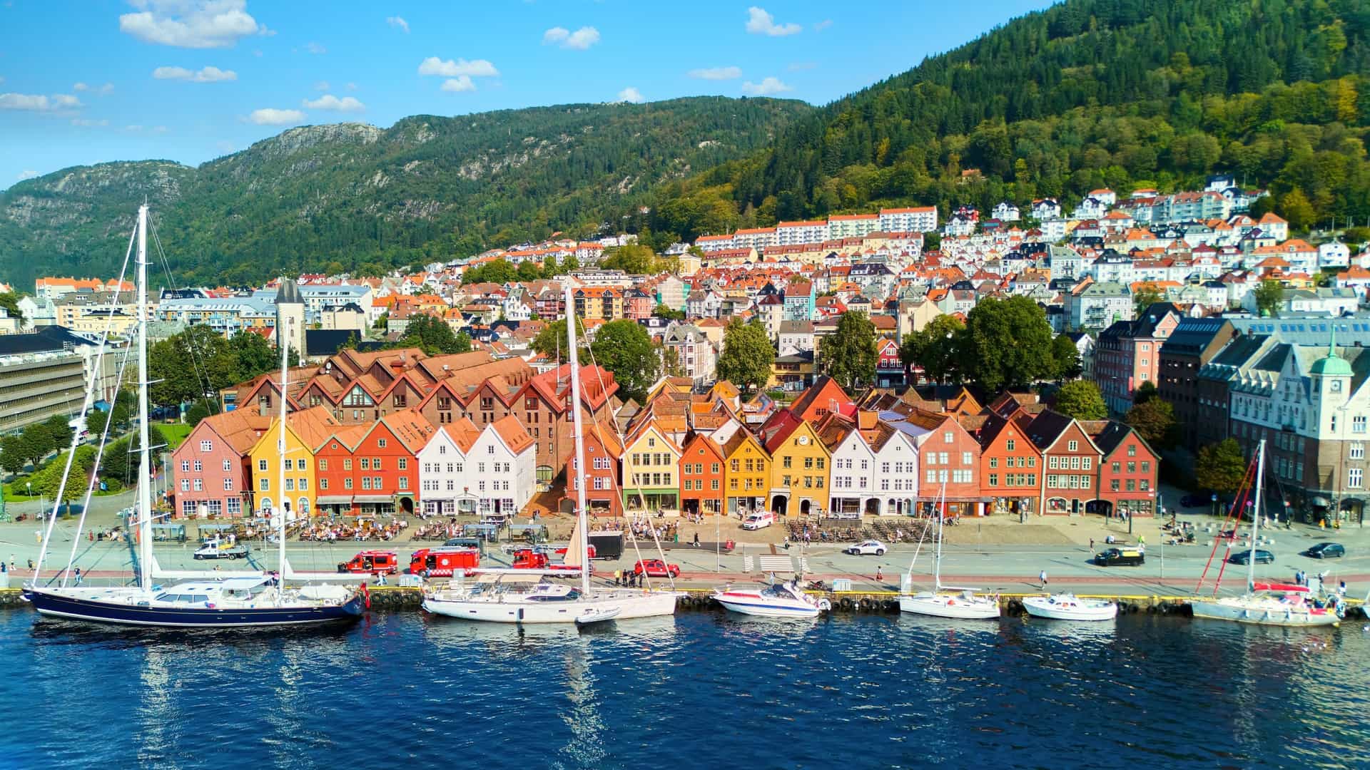 An aerial view of the historic Bryggen district in Bergen, Norway. The image shows the iconic rows of colorful wooden buildings with their pointed gables facing the Vågen harbor. The water in the foreground reflects the buildings and the surrounding mountains are visible in the background.