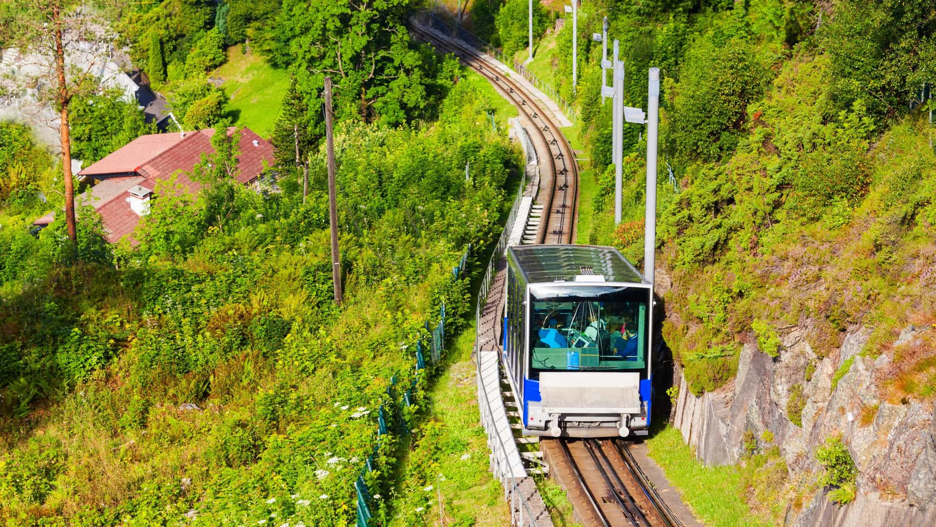 A scenic view of the Fløibanen funicular railway in Bergen, Norway, as it travels up the steep tracks of Mount Fløyen. The funicular car is visible on the track, which is surrounded by lush green trees and rocky terrain. The city of Bergen and its harbor are visible in the background, under a clear sky.