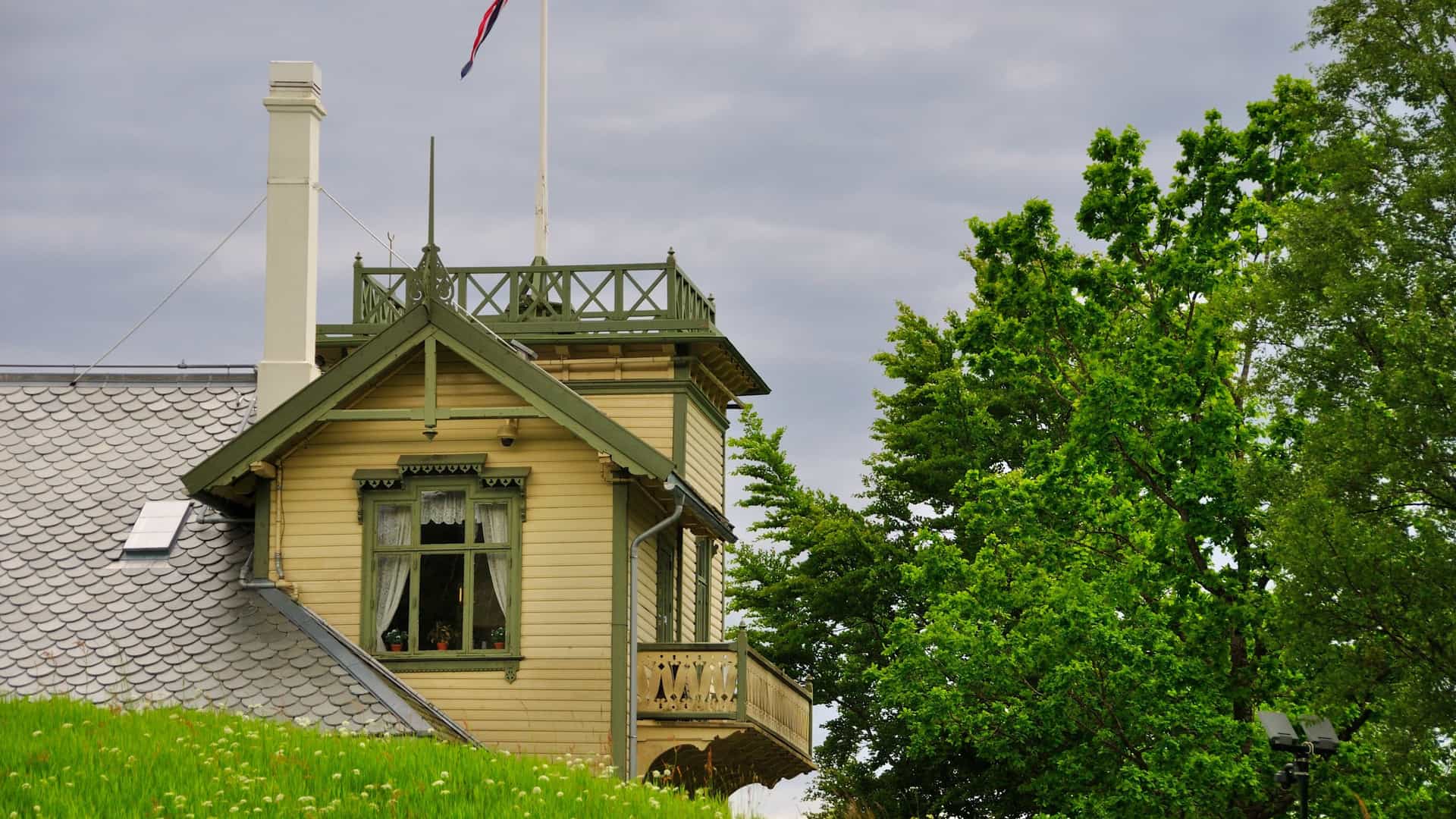 A serene view of Troldhaugen, the former home and museum of Norwegian composer Edvard Grieg, located near Bergen, Norway. The image shows the yellow, two story wooden villa built in a Swiss chalet style, set amidst a lush green lawn and trees. In the foreground, Grieg's small, rustic, lakeside Composer's Hut is visible, situated right at the edge of the water overlooking Nordås Lake. The peaceful setting highlights the inspirational landscape where Grieg composed many of his most famous works.