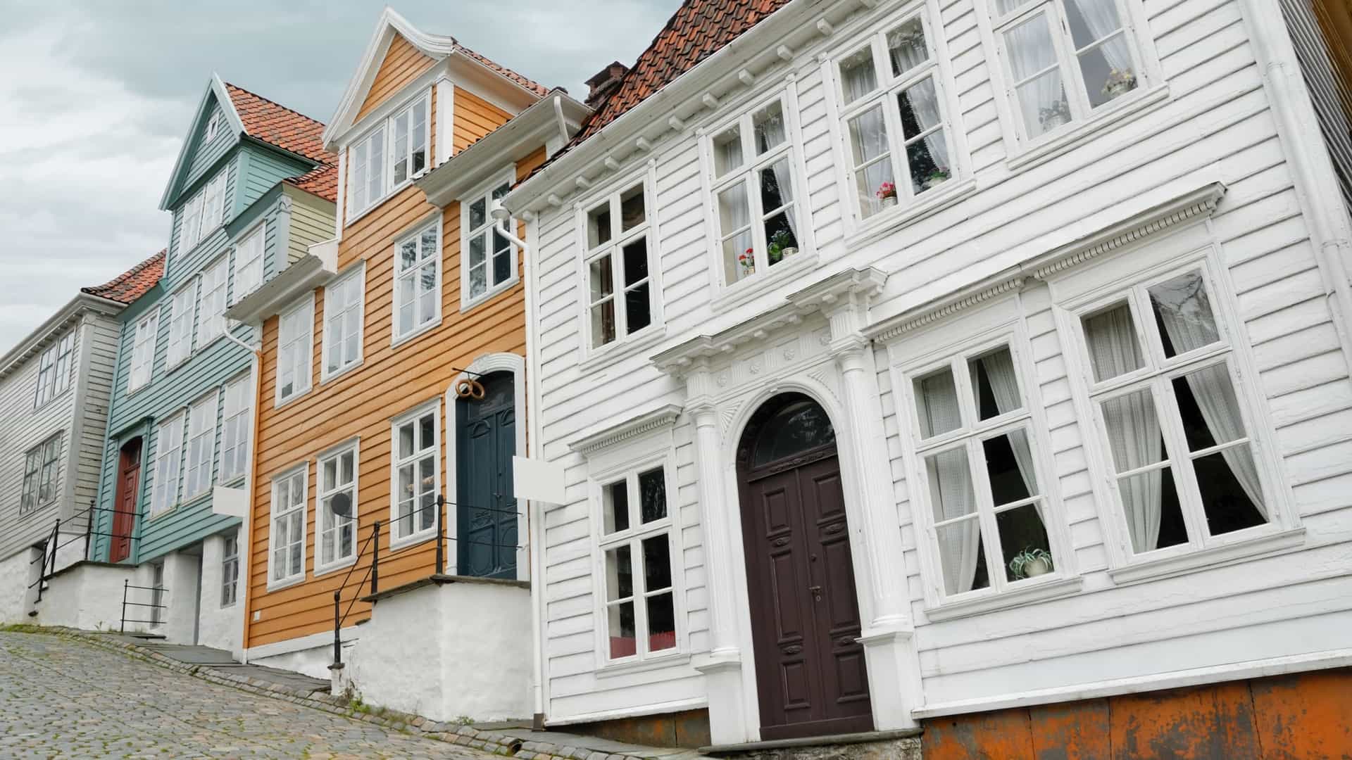 A close up detail of the second story facade of a traditional 18th or 19th century wooden house at the Gamle Bergen (Old Bergen) open air museum in Bergen, Norway, featuring white clapboard siding, a balcony with a simple wooden railing, and open windows with decorative trim.