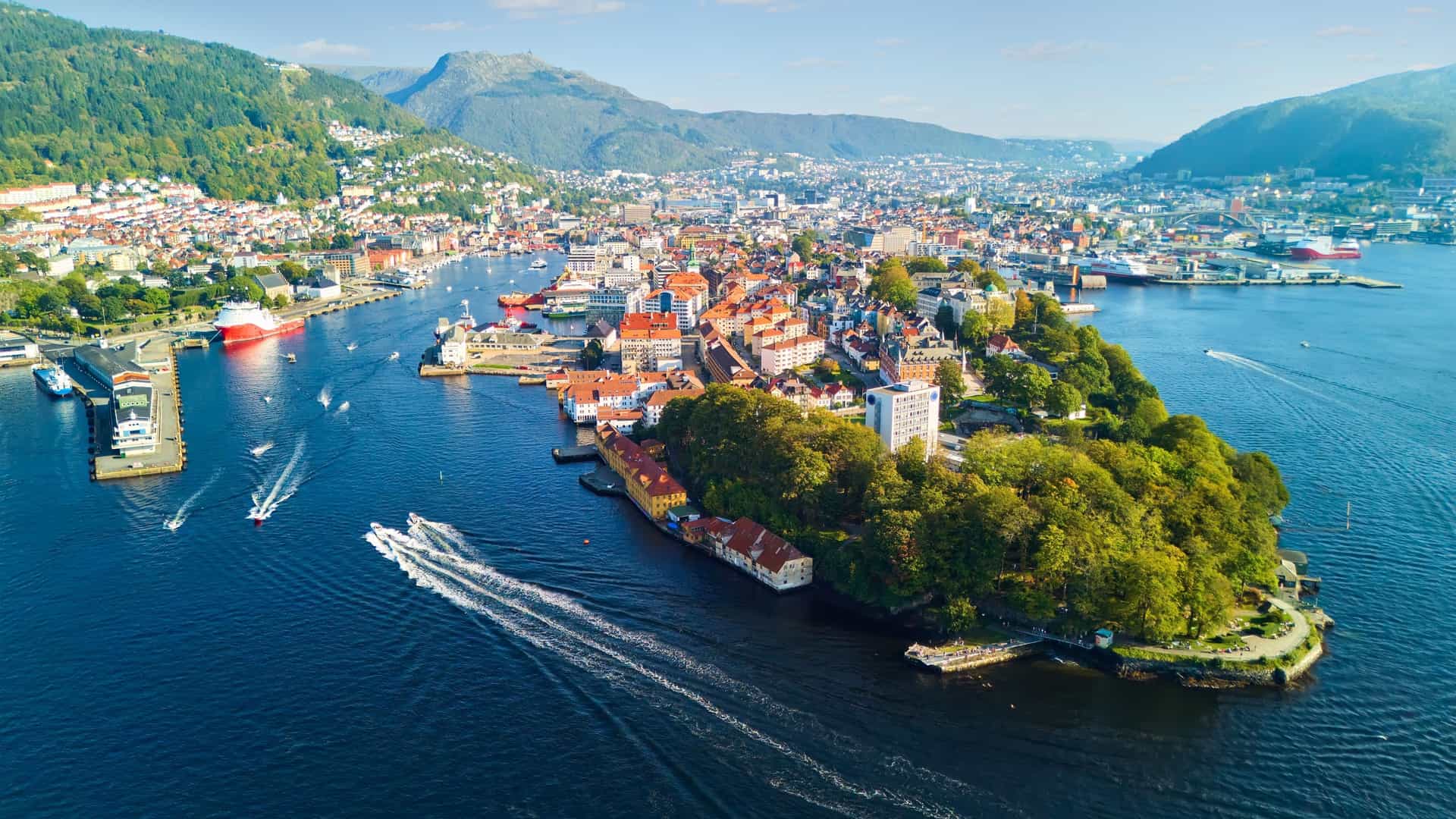 An aerial view of Bergen, Norway, showing the dense collection of buildings and streets of the old town on the Nordnes peninsula. The peninsula is surrounded by water, with boats visible in the harbor, and is set against a backdrop of mountains. The image is a high angle shot, capturing the full extent of the peninsula and the surrounding cityscape.