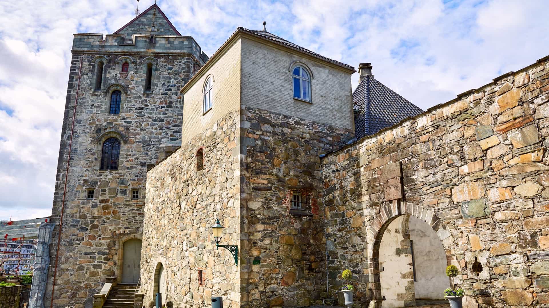 A scenic view of the historic Bergenhus Fortress in Bergen, Norway. The image shows several stone buildings and towers of the fortress complex, including the Rosenkrantz Tower, with the city's harbor and surrounding mountains in the background. The sky is bright and clear, highlighting the stone walls and architecture of the fortress.