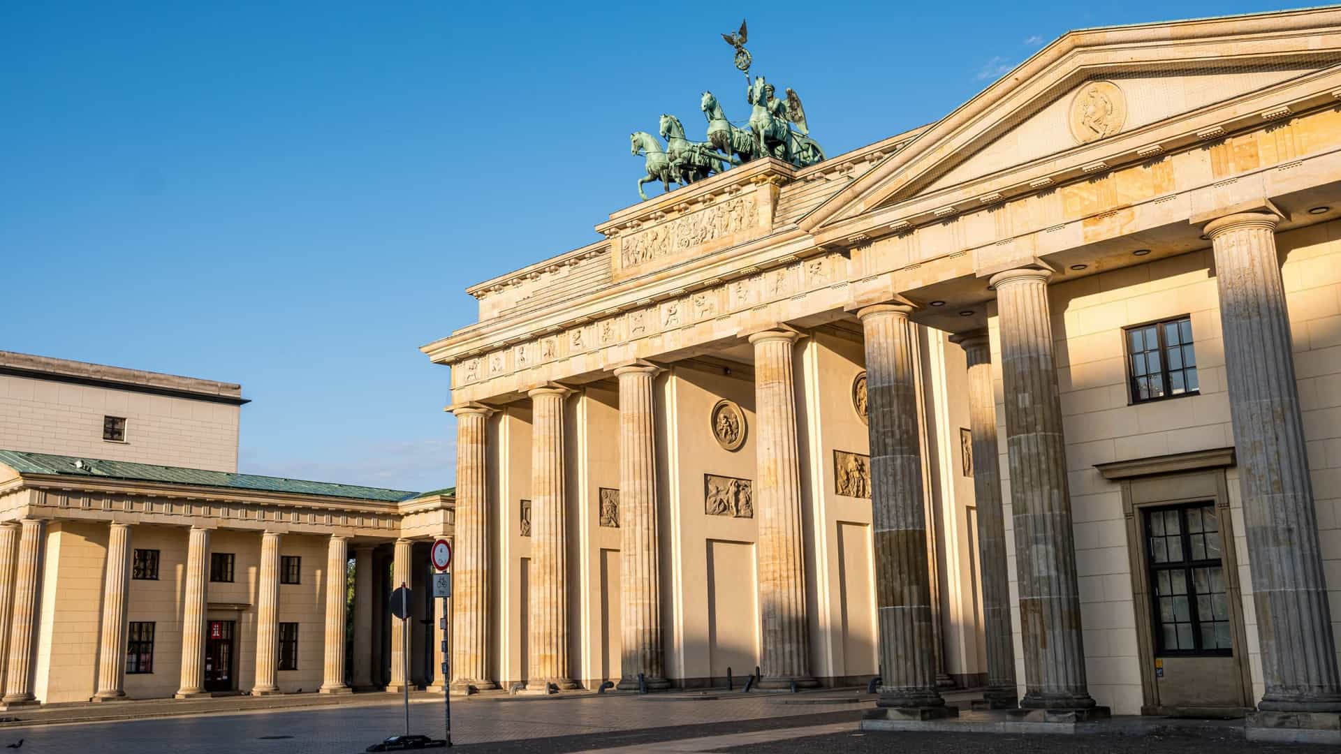 The iconic Brandenburg Gate in Berlin, a neoclassical monument with twelve Doric columns and the Quadriga statue on its roof, is presented from a head on perspective on a bright, sunny day. The large, open cobblestone area in front of the gate is clear, and the sky above is a clear blue.