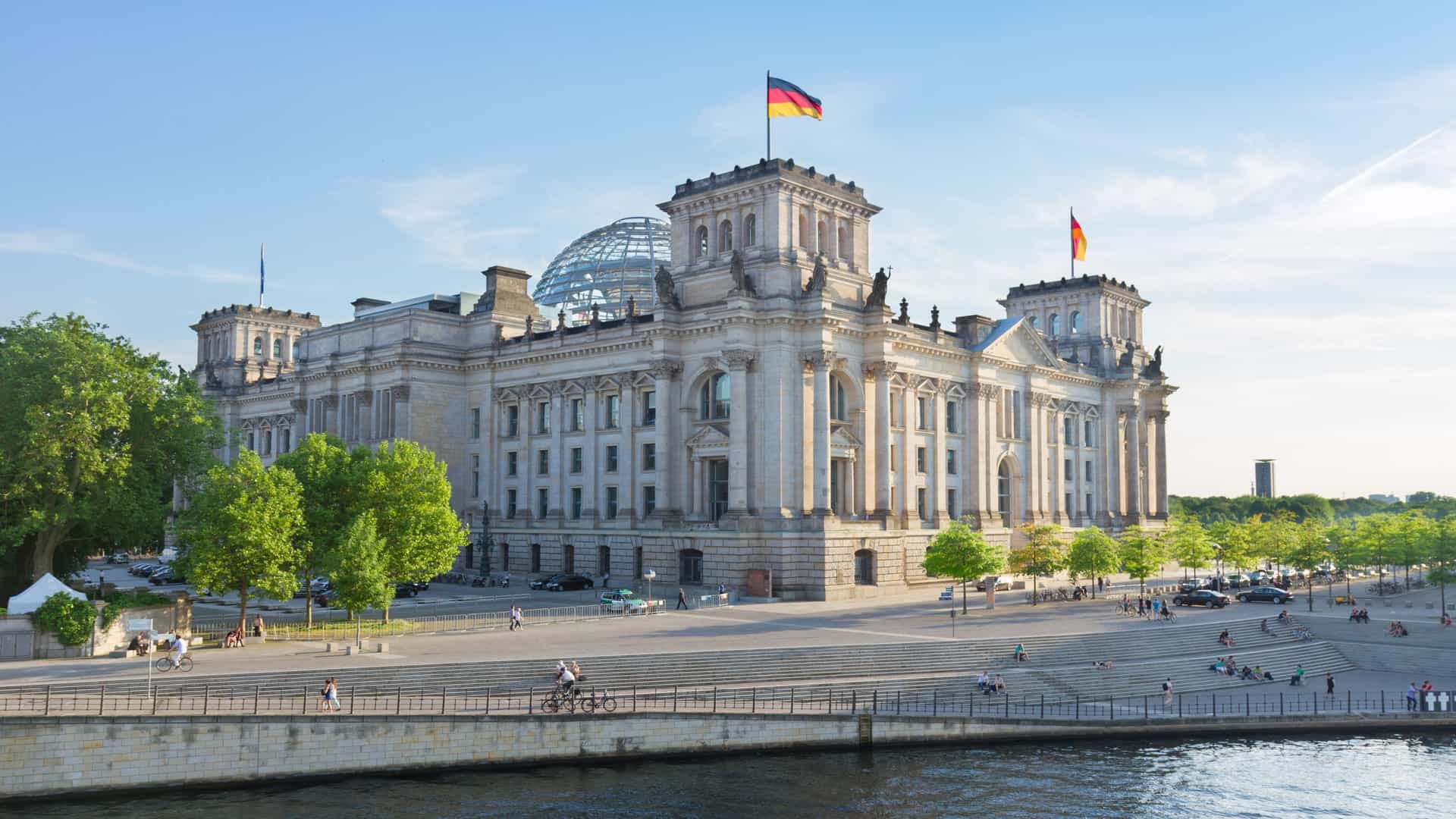A scenic view of the Reichstag building, the seat of the German parliament, located on the banks of the River Spree in Berlin. The historic building, with its modern glass dome, is reflected in the calm water of the river on a clear day with a blue sky.